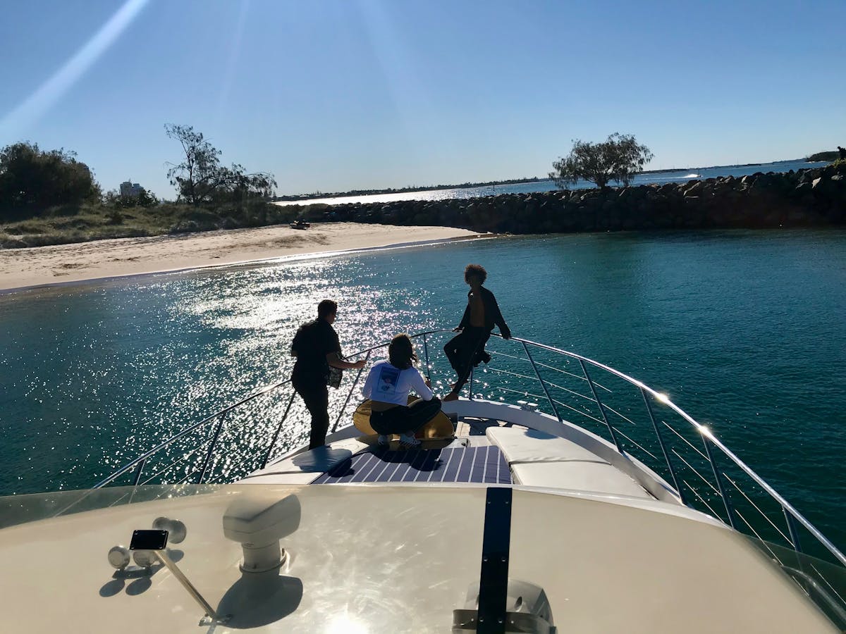 Three people enjoying the bow of a private yacht anchored near a sandy beach on the Gold Coast.