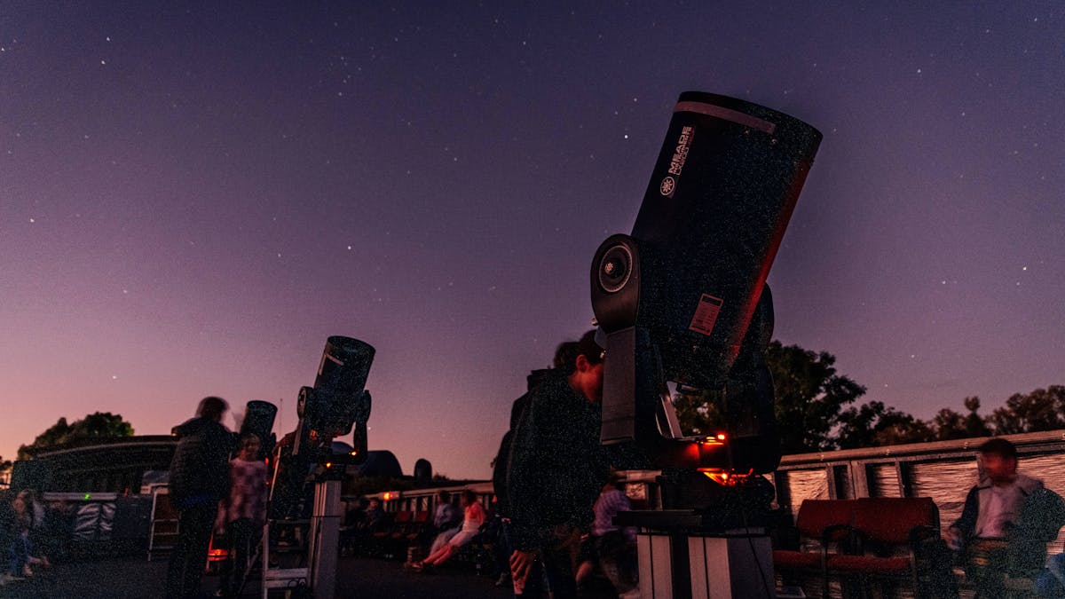 Multiple telescopes pointing up, with guests looking through the telescopes. Twilight sky behind.