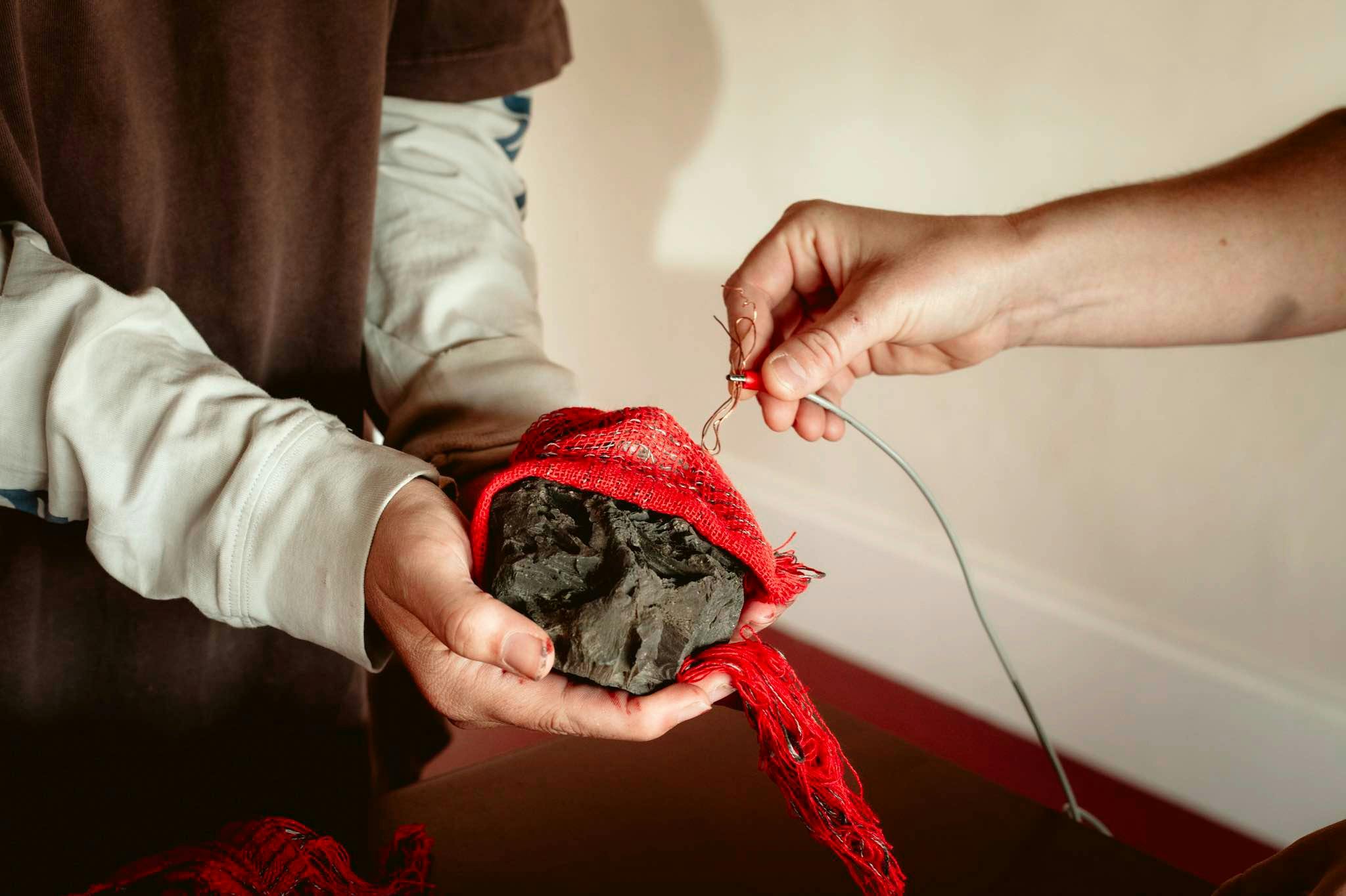 A person holds a rock wrapped in red fabric, another person holds an electrical wire to the rock