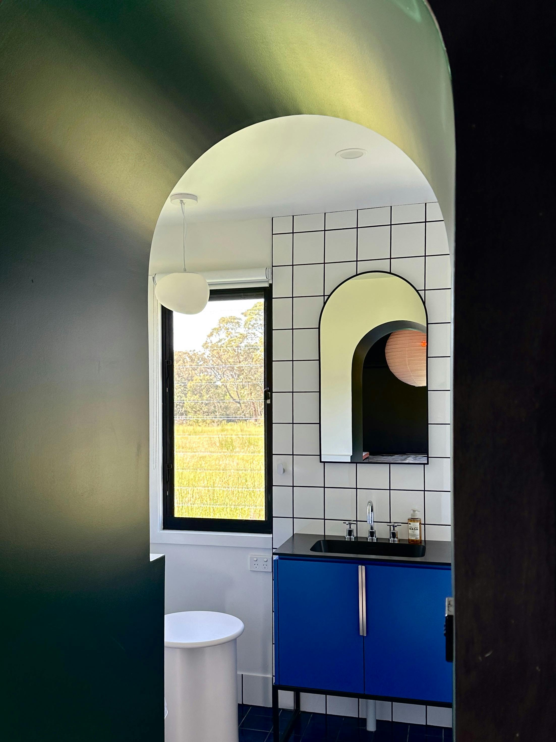 Arched doorway leading into a contemporary white tiled bathroom with blue vanuty and louvre windows