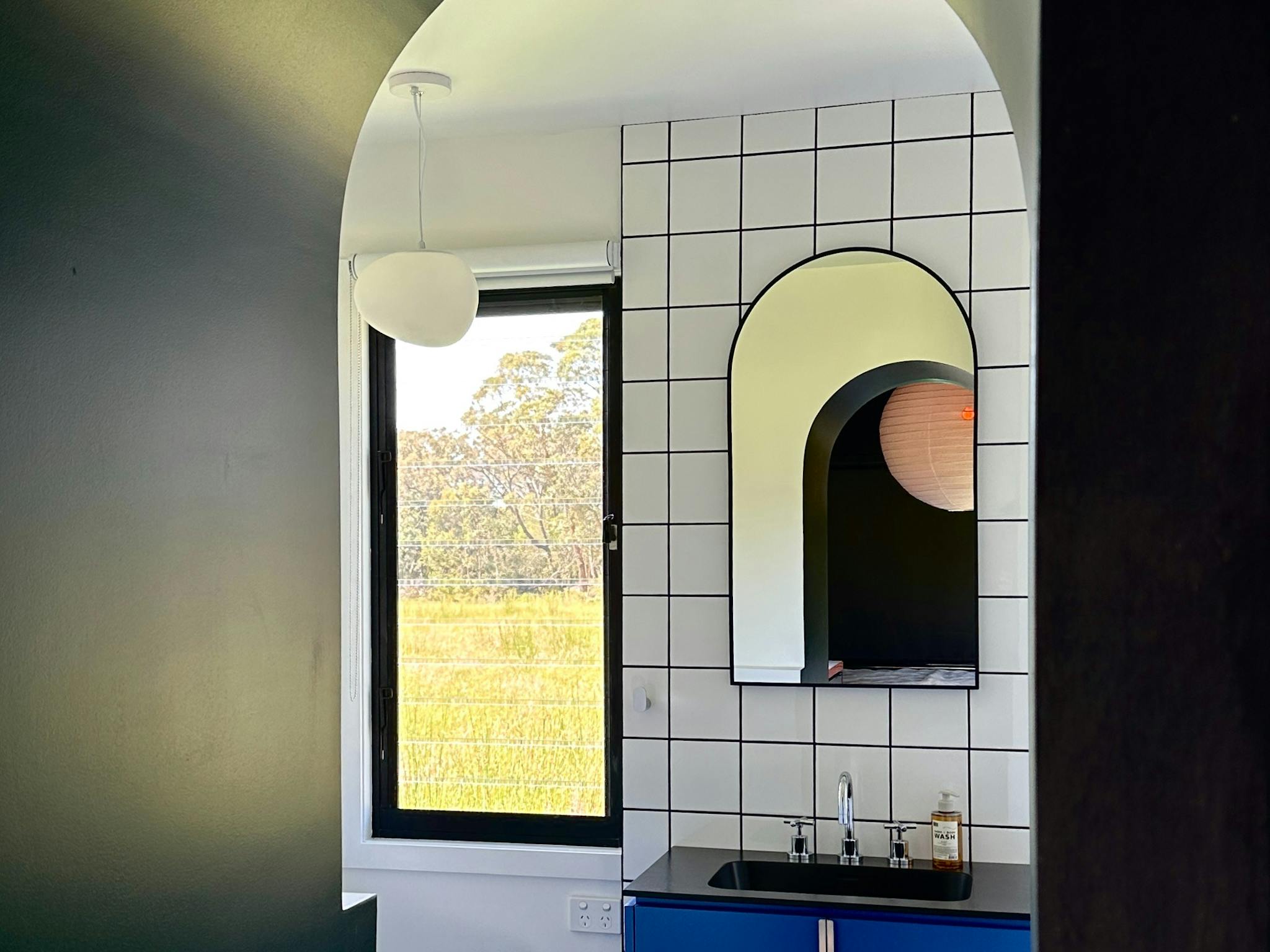 Arched doorway leading into a contemporary white tiled bathroom with blue vanuty and louvre windows