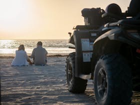 Couple sitting on the beach watching sunset with quad bike atv out of focus in foreground
