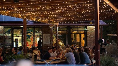 People sitting along outdoor tables in the evening time, underneath fairy lights