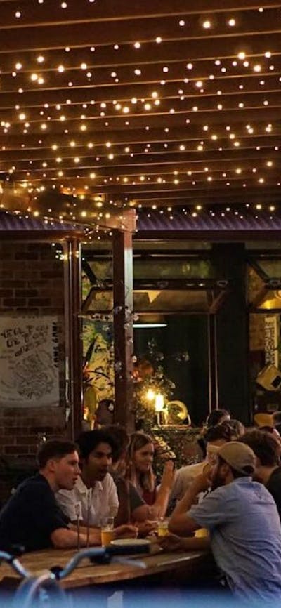 People sitting along outdoor tables in the evening time, underneath fairy lights