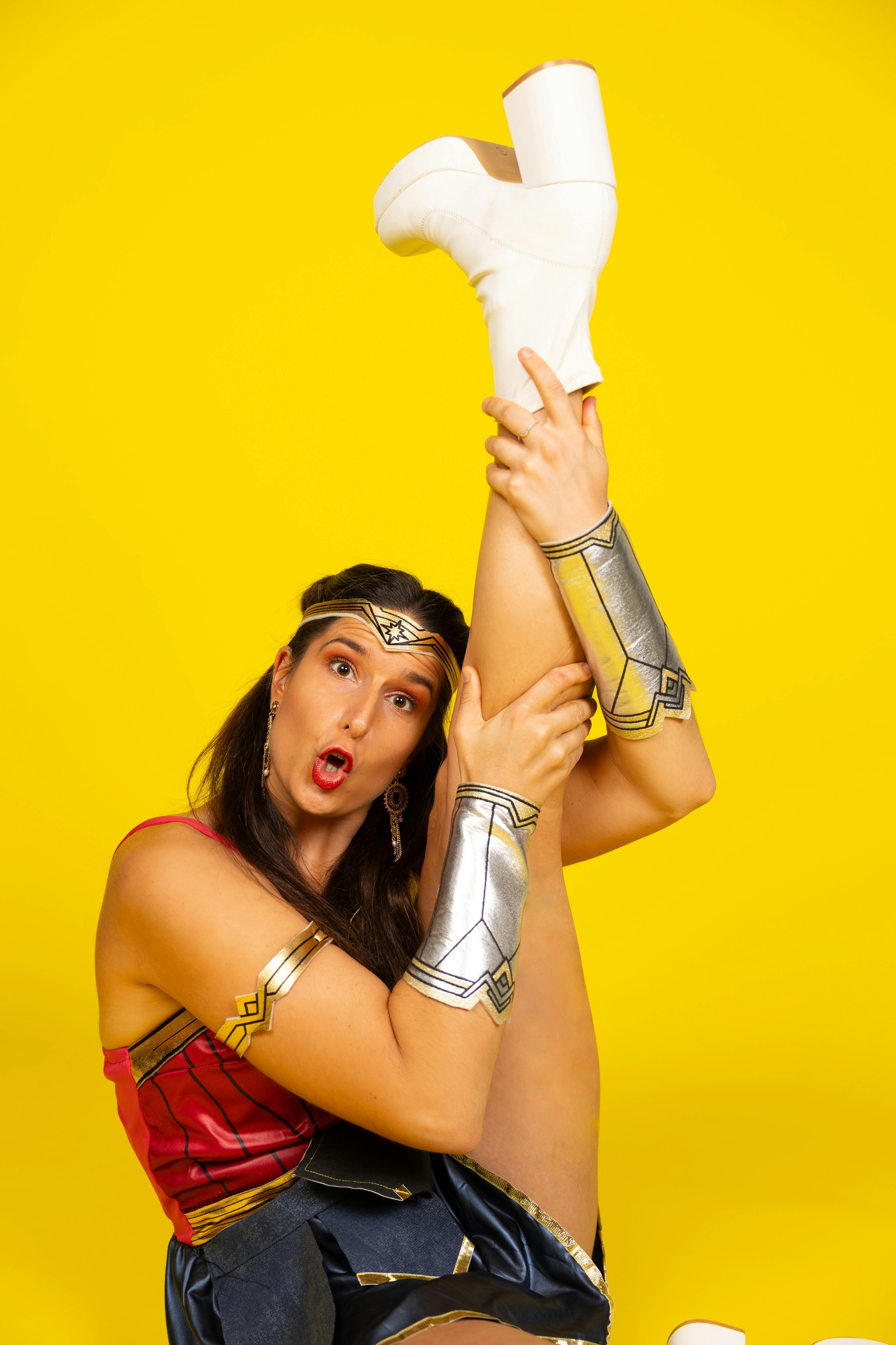 An expressive woman holding her leg in the air, dressed as Wonder Woman on a yellow background.