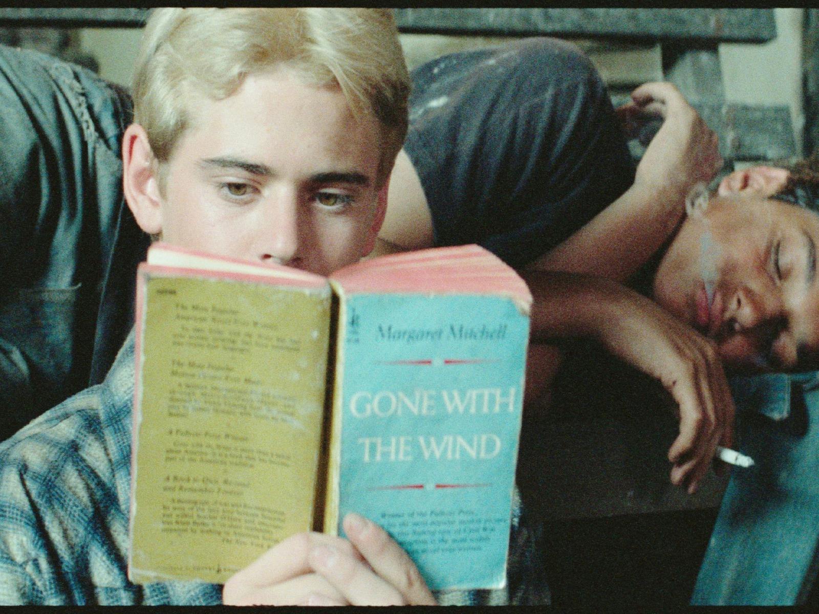 One male lying on couch sleeping, another sitting in front reading a book