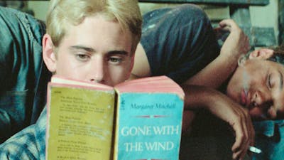 One male lying on couch sleeping, another sitting in front reading a book