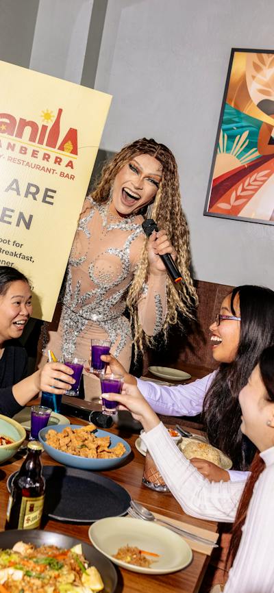 A performer holding a microphone stands beside a table where guests raise purple drinks in a toast,