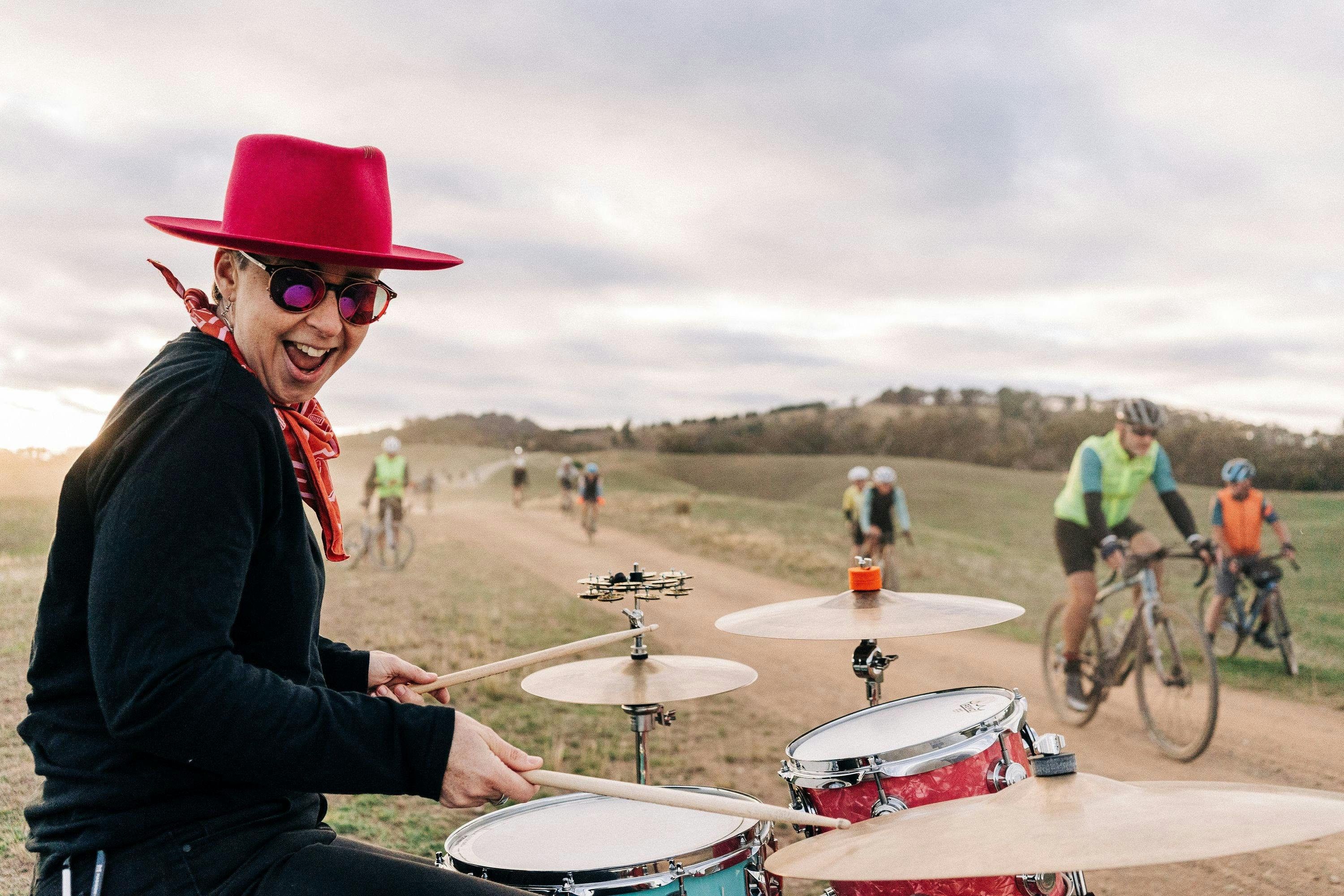 Drummer smiling at the camera while cyclists ride past on gravel road.