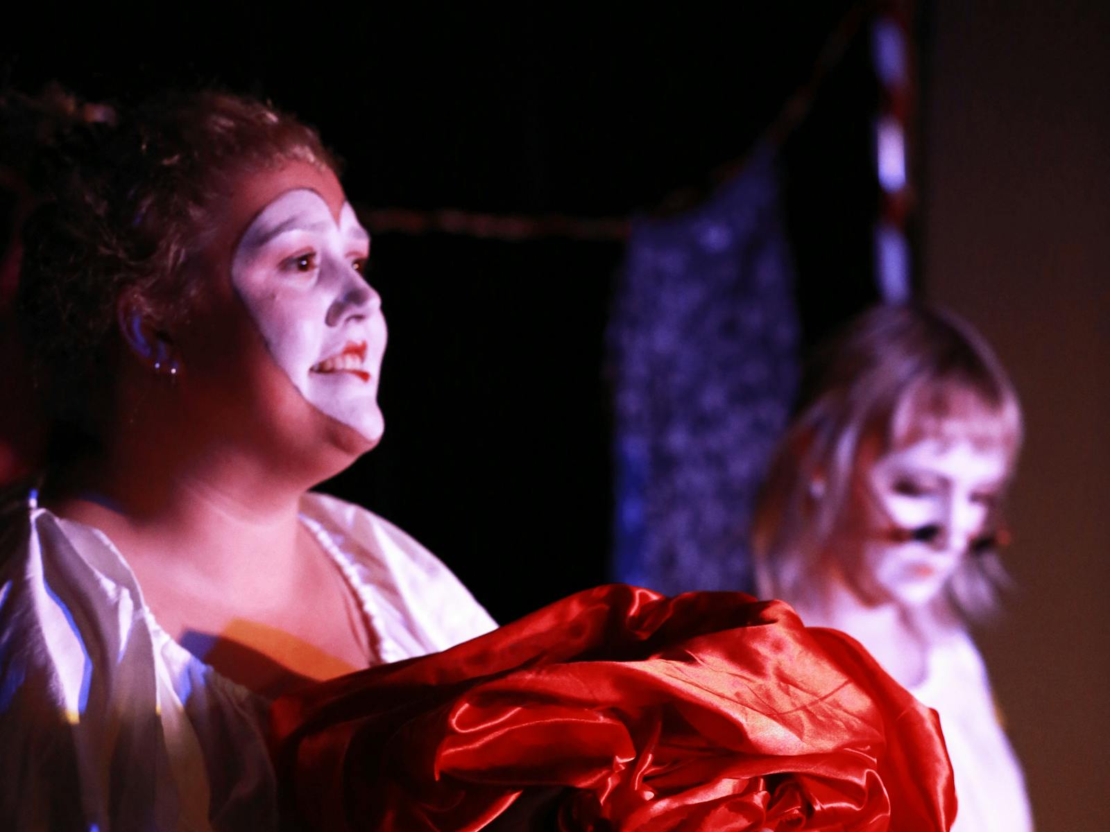A performer in white with a white painted face sings out to the audience while clutching red fabric.