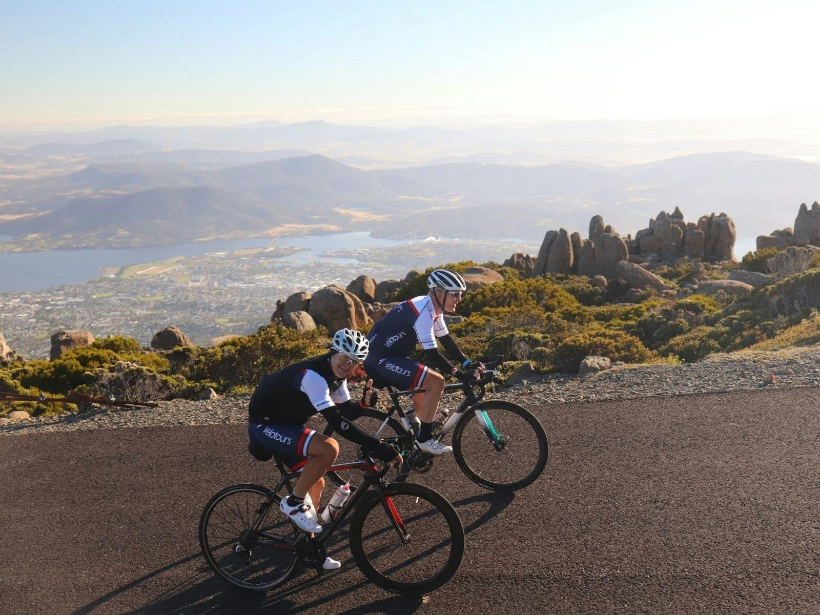 men on bikes looking at camera as they ride to top of mountain with city backdrop in background