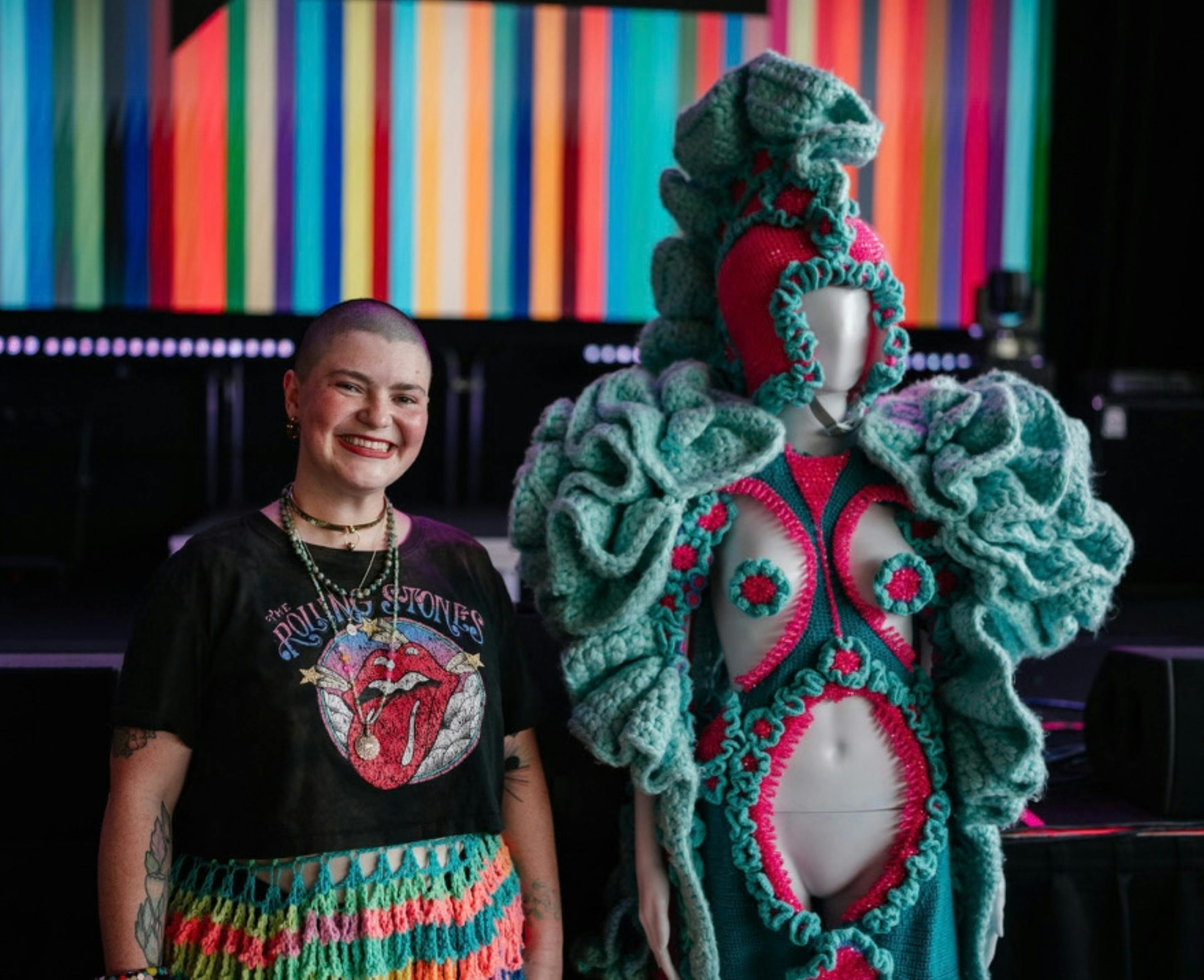 A woman standing next to handmade wearable yarn art.