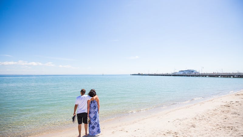 couple enjoy a walk along the beach. Clean blue water, white sand and the pier in the foreground.