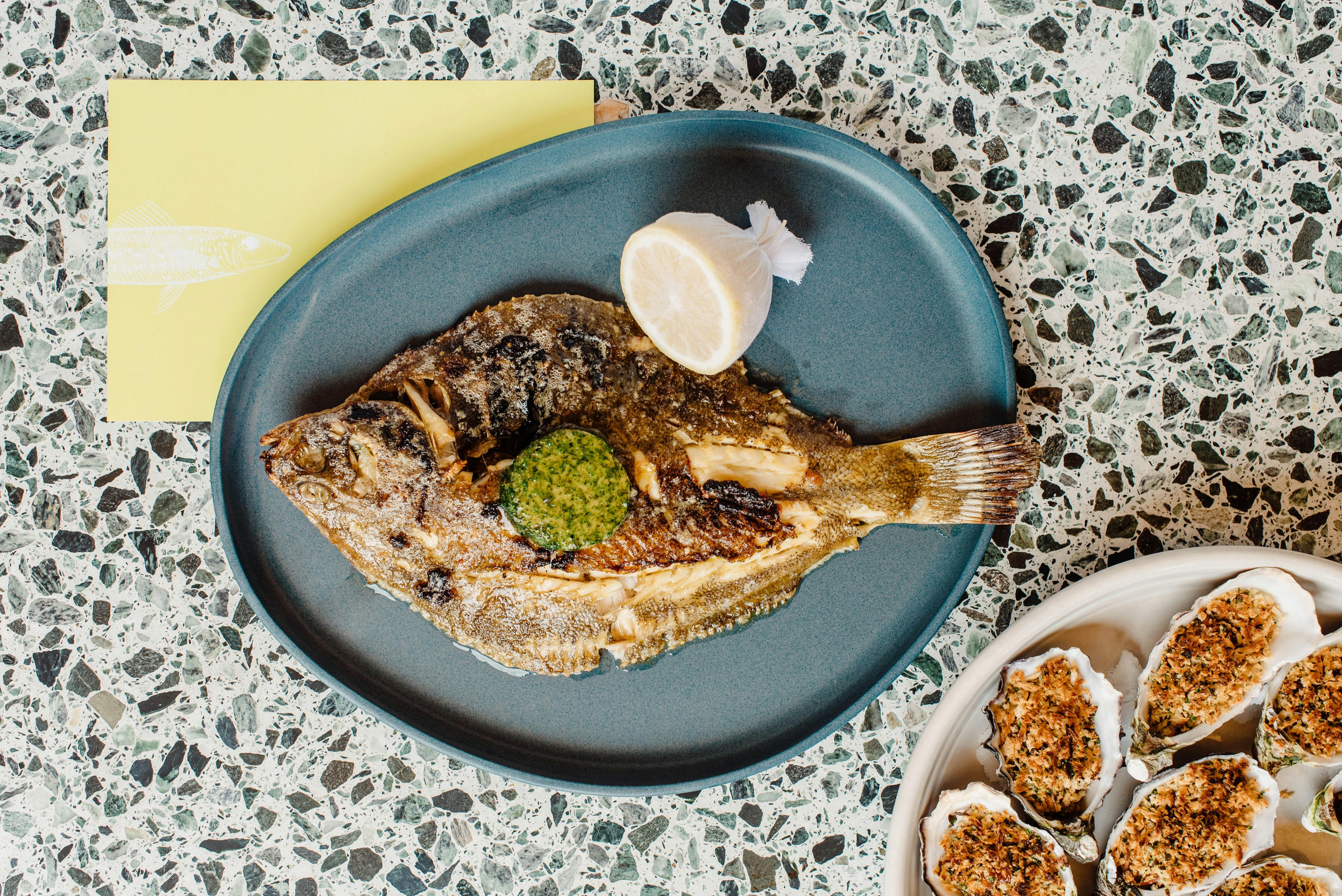 A large grilled fish on a blue plate, with another plate of cooked oyster to the side