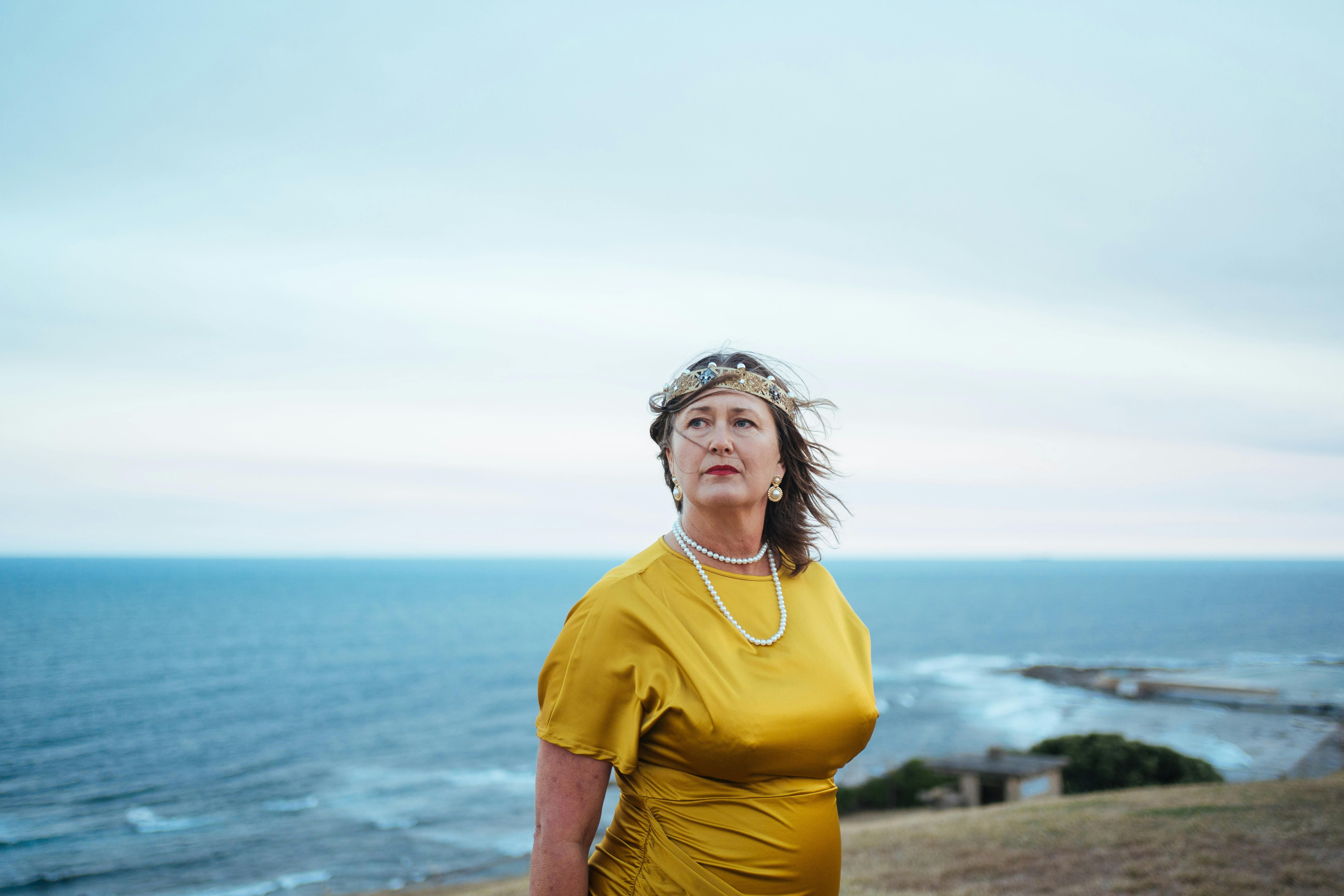 This photo pictures a woman in a gold dress standing against the backdrop of Newcastle's coastline