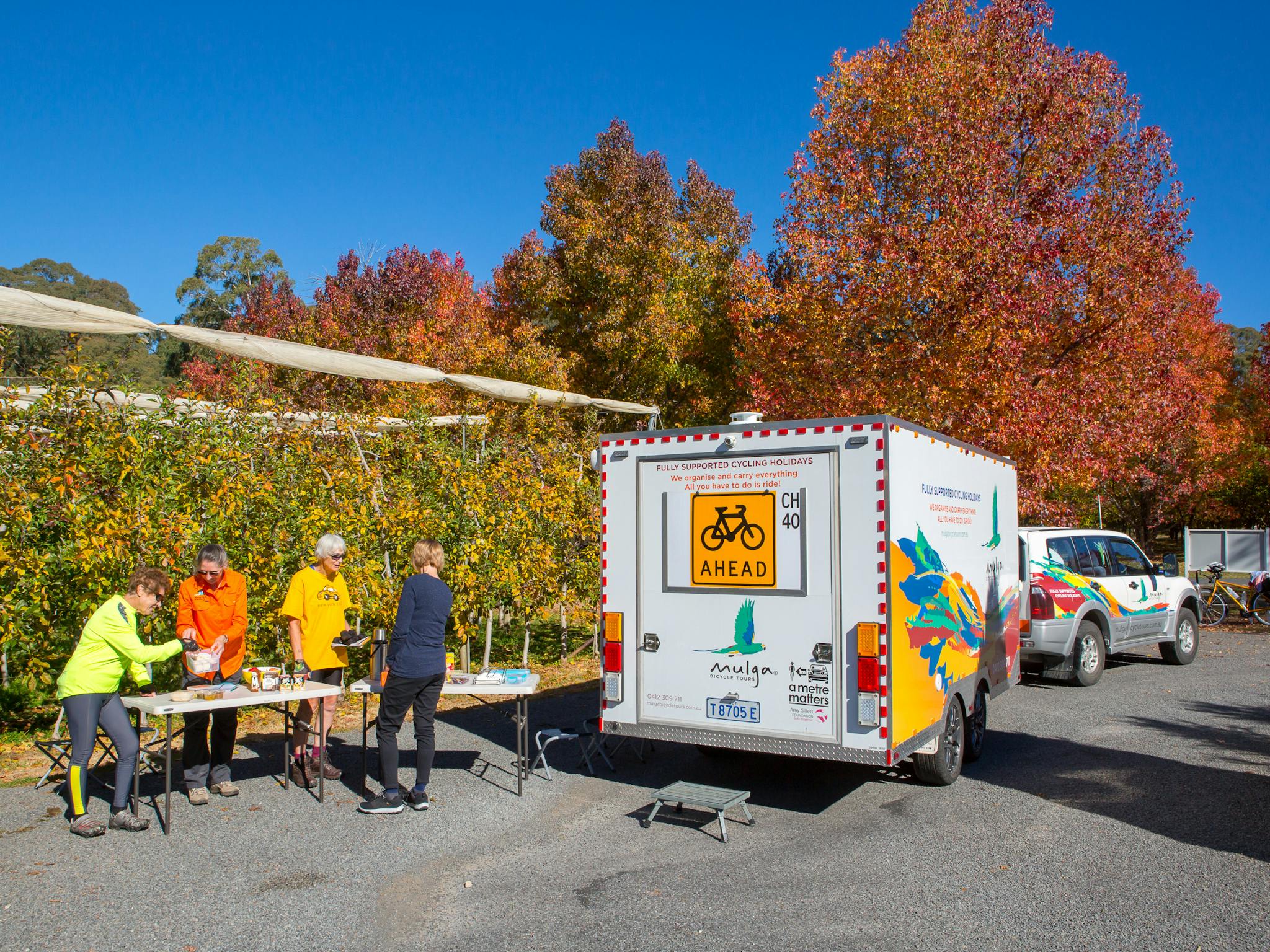 Guests take refreshments from the Mulga support trailer next to an orchard in full autumn colours