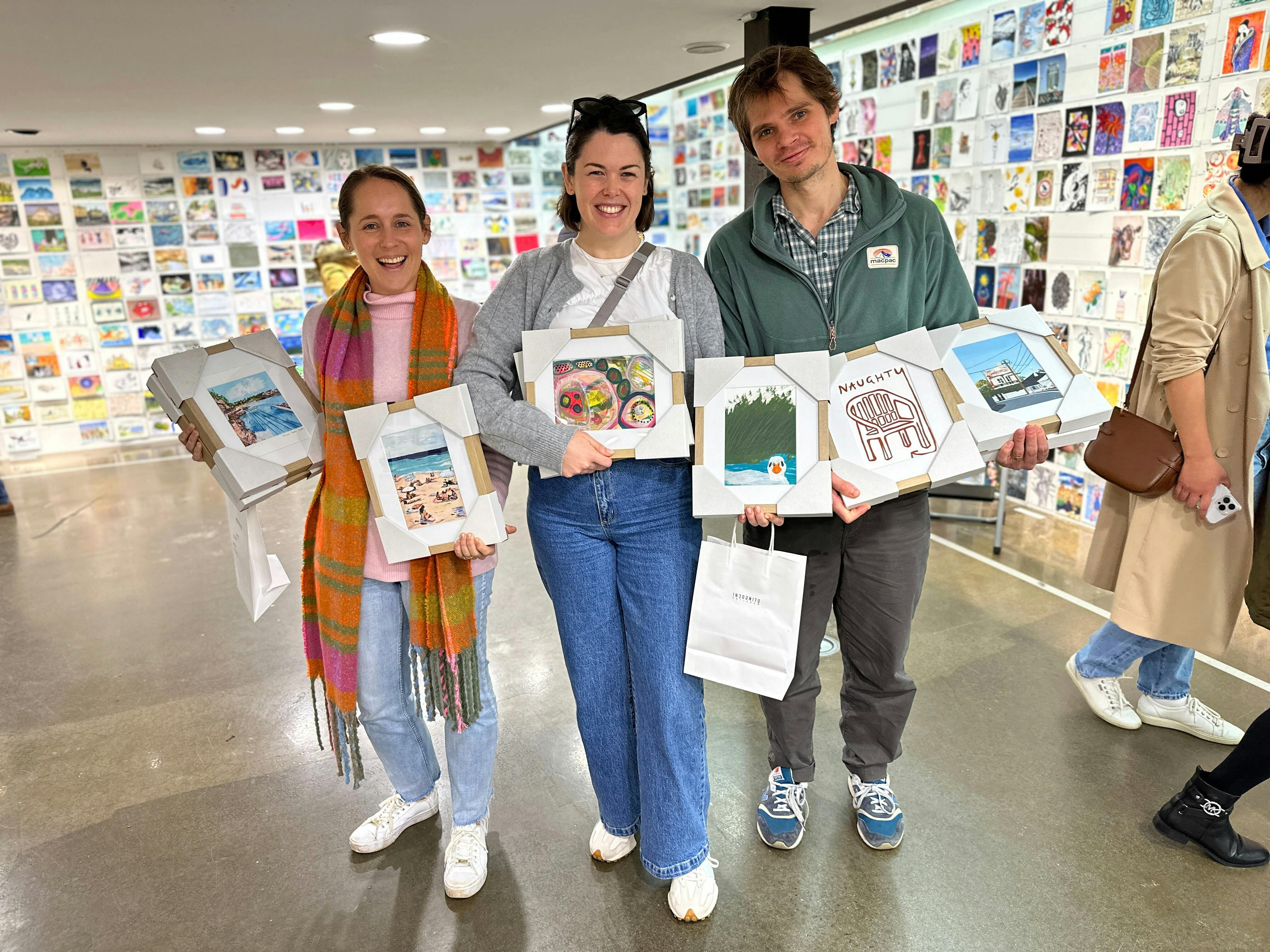 Three people holding framed A5 original artworks they bought at the Incognito Art Show.
