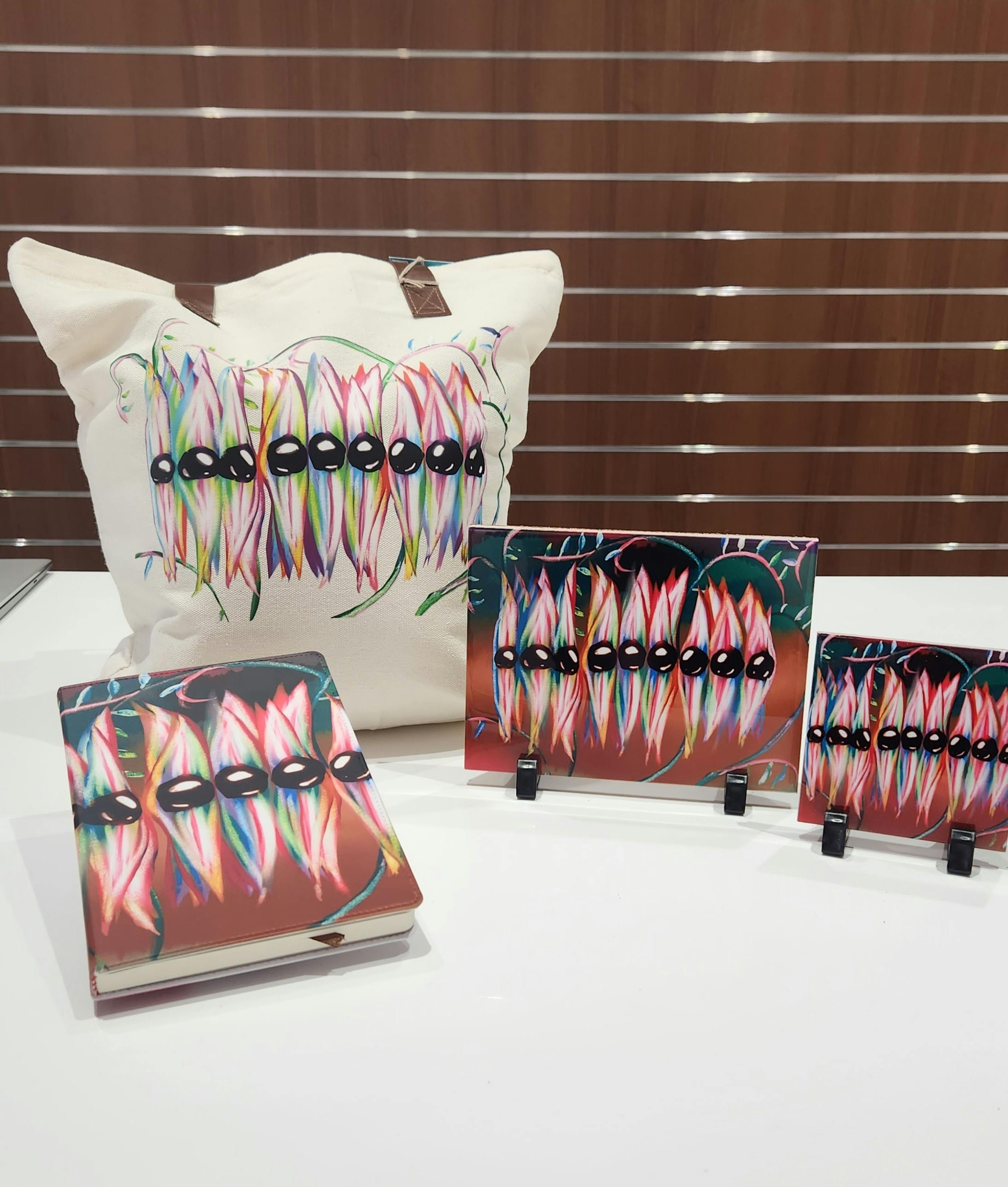 A display of tote bag, journal, coast with Sturt's desert peas designs on a white counter