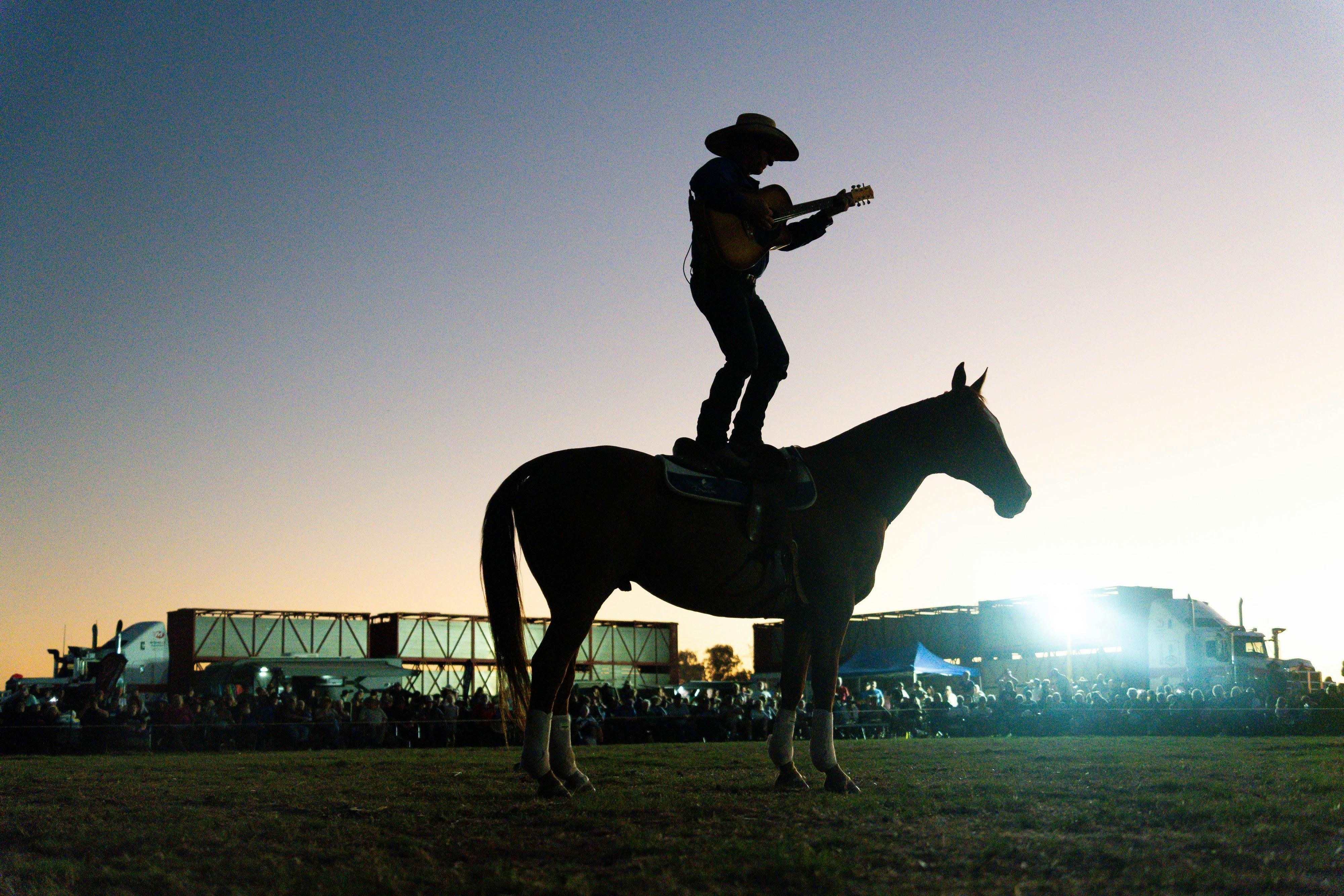 Tom Curtain standing on a horse while playing guitar and singing a song