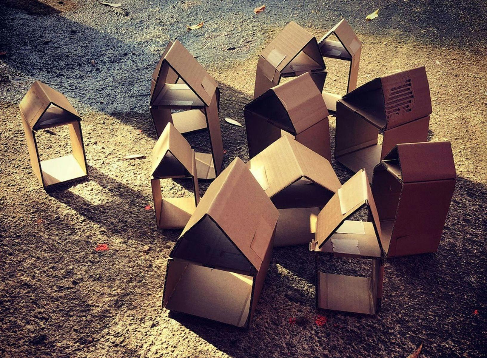 A cluster of small cardboard houses, in sunlight on a gravel surface.