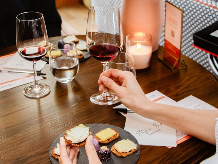 Hand holding a cracker with cheese and another hand holding a wine glass.