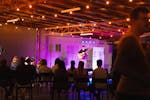 A musician performs on stage in front of an audience in an industrial space with string lights.