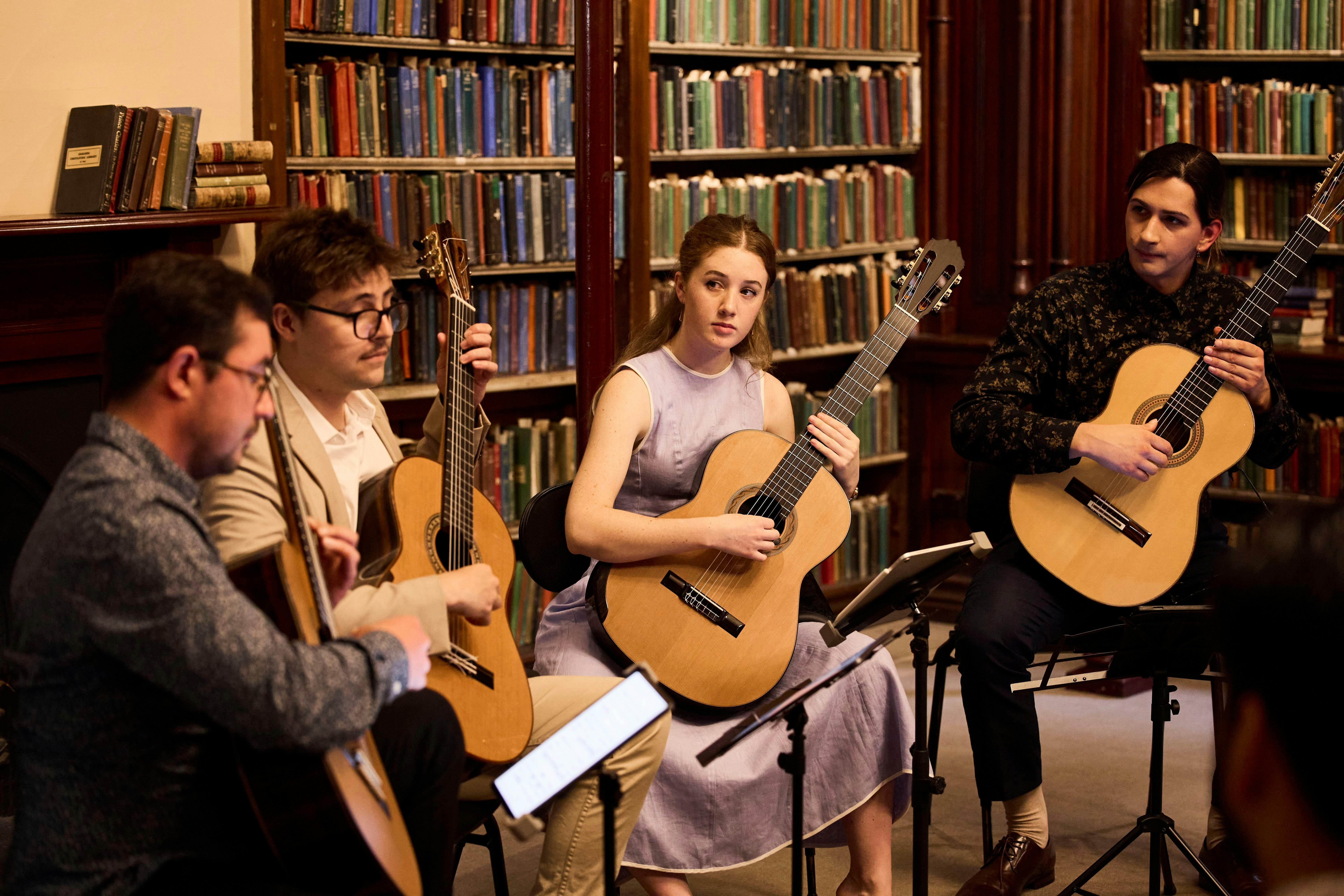 A guitar quartet perform in a library space with shelves full of books behind them.