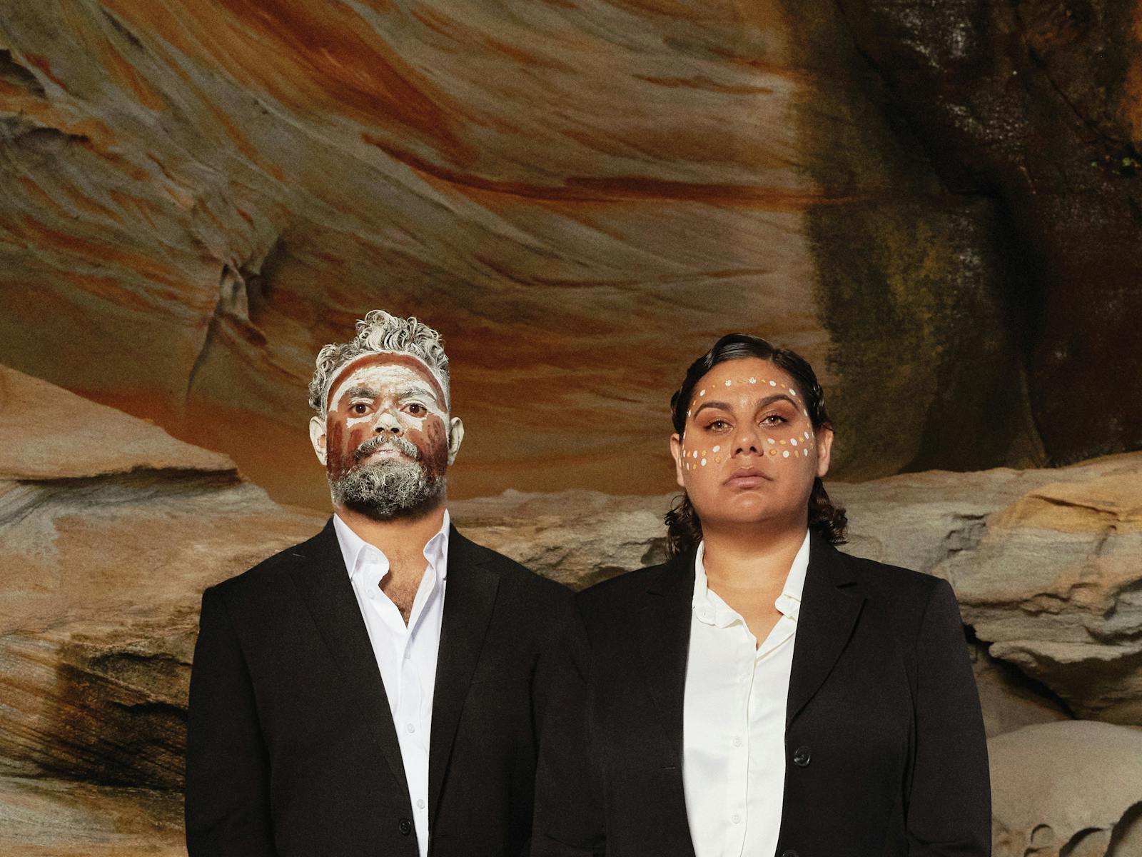 Two first nations people, are dressed in formal western suits, with their faces painted in ochre.