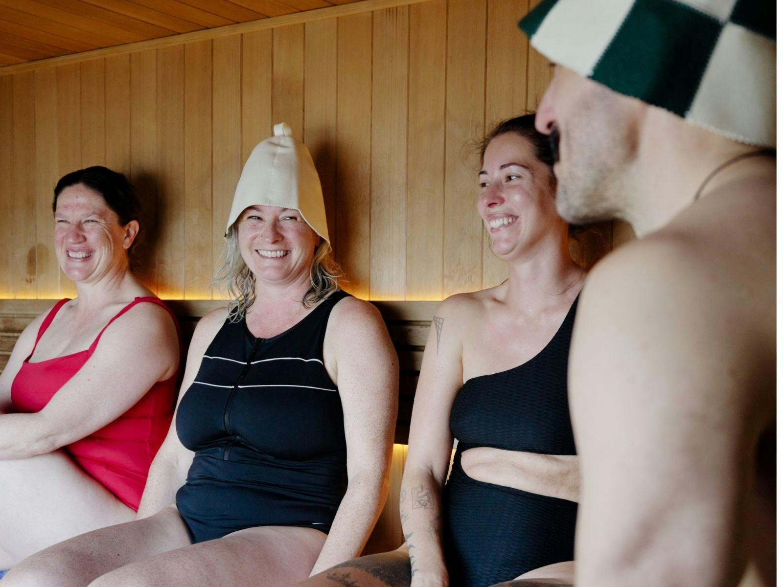A group of smiling people sitting in a traditional sauna with sauna hats on