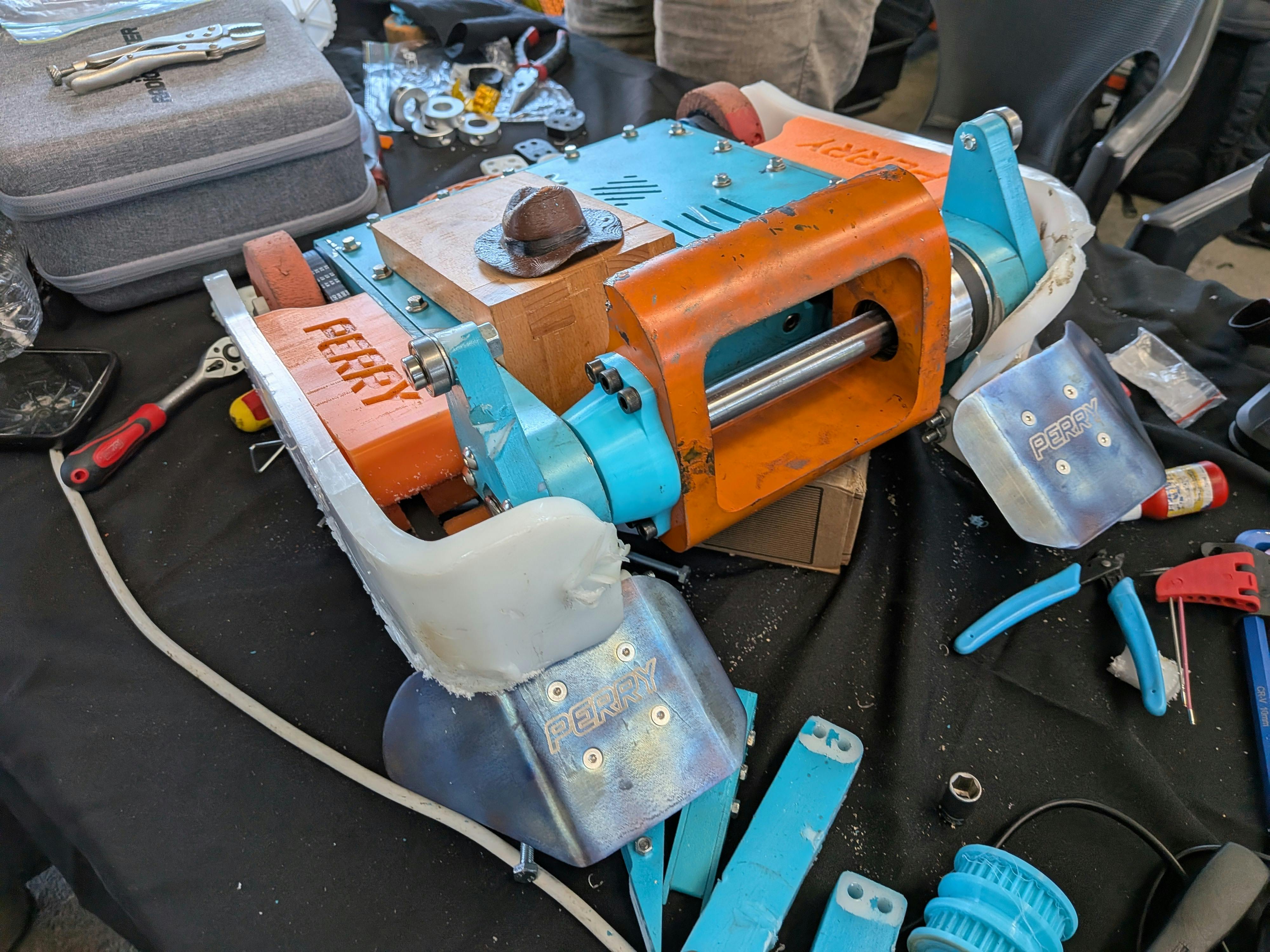Turquoise and orange combat robot with a drum weapon on a workbench in the Robowars pits.