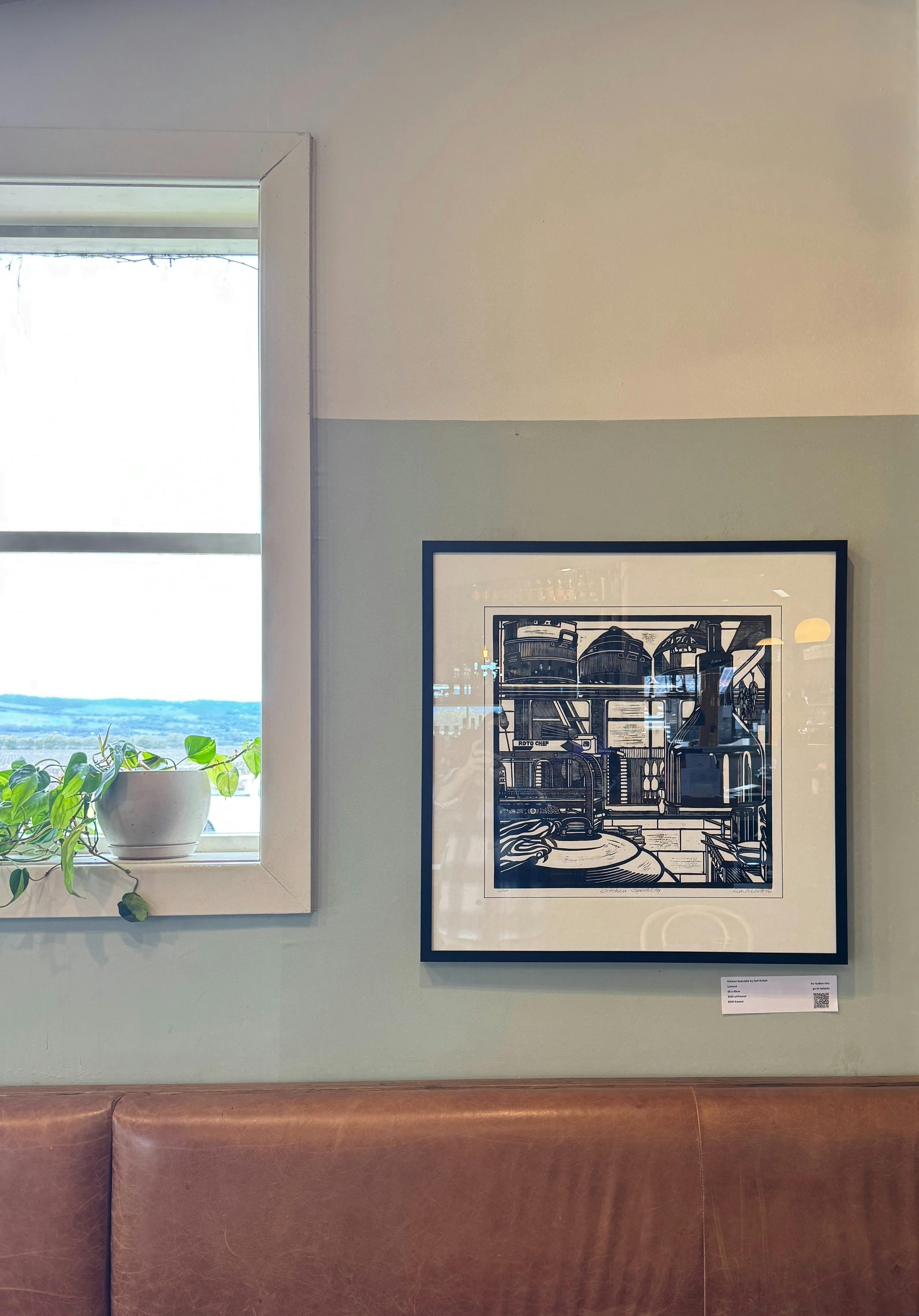 A linocaut framed picture on a green wall next to a window with a plant on the shelf.