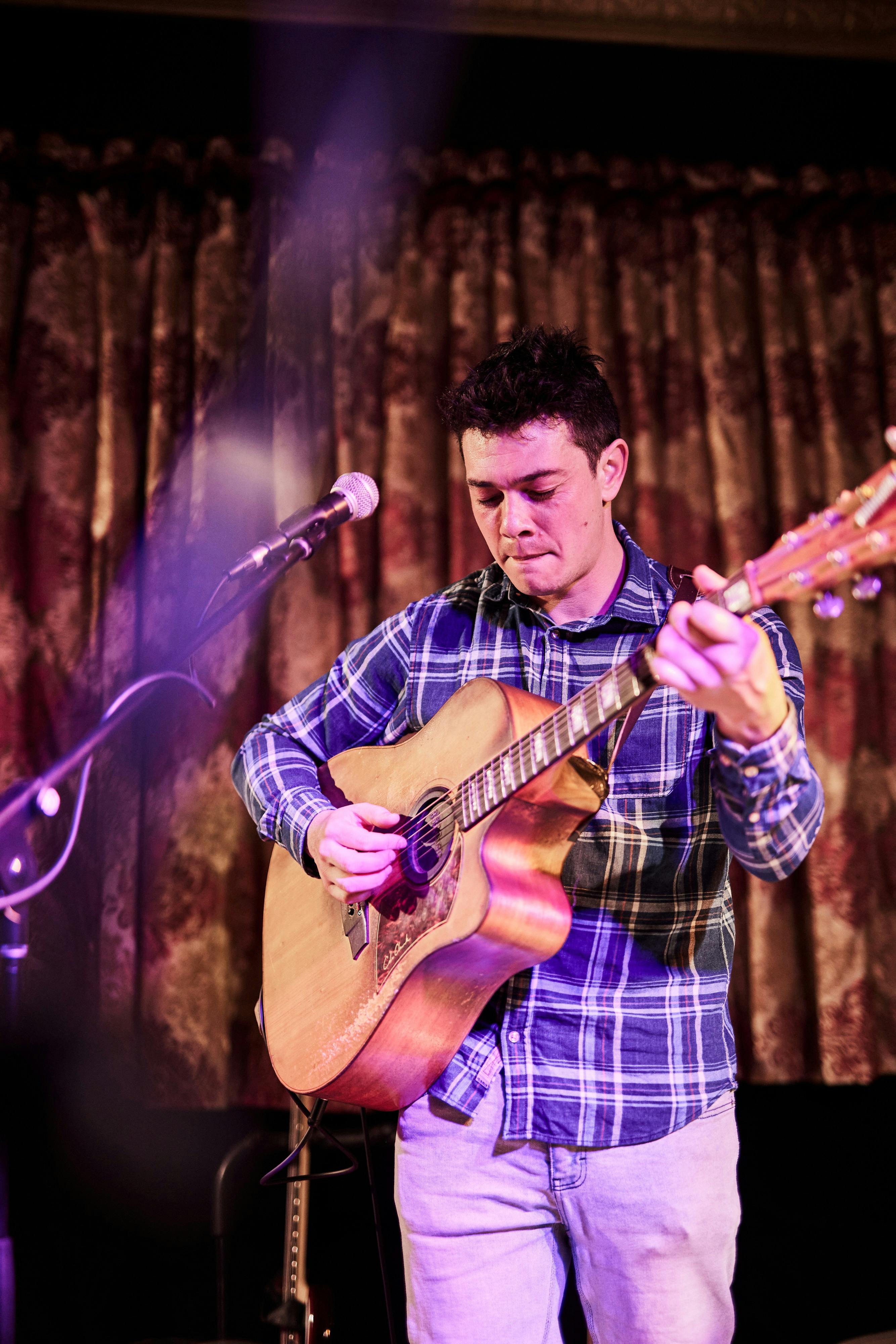 A musician plays guitar standing in front of a microphone,  with damasque curtains in the background