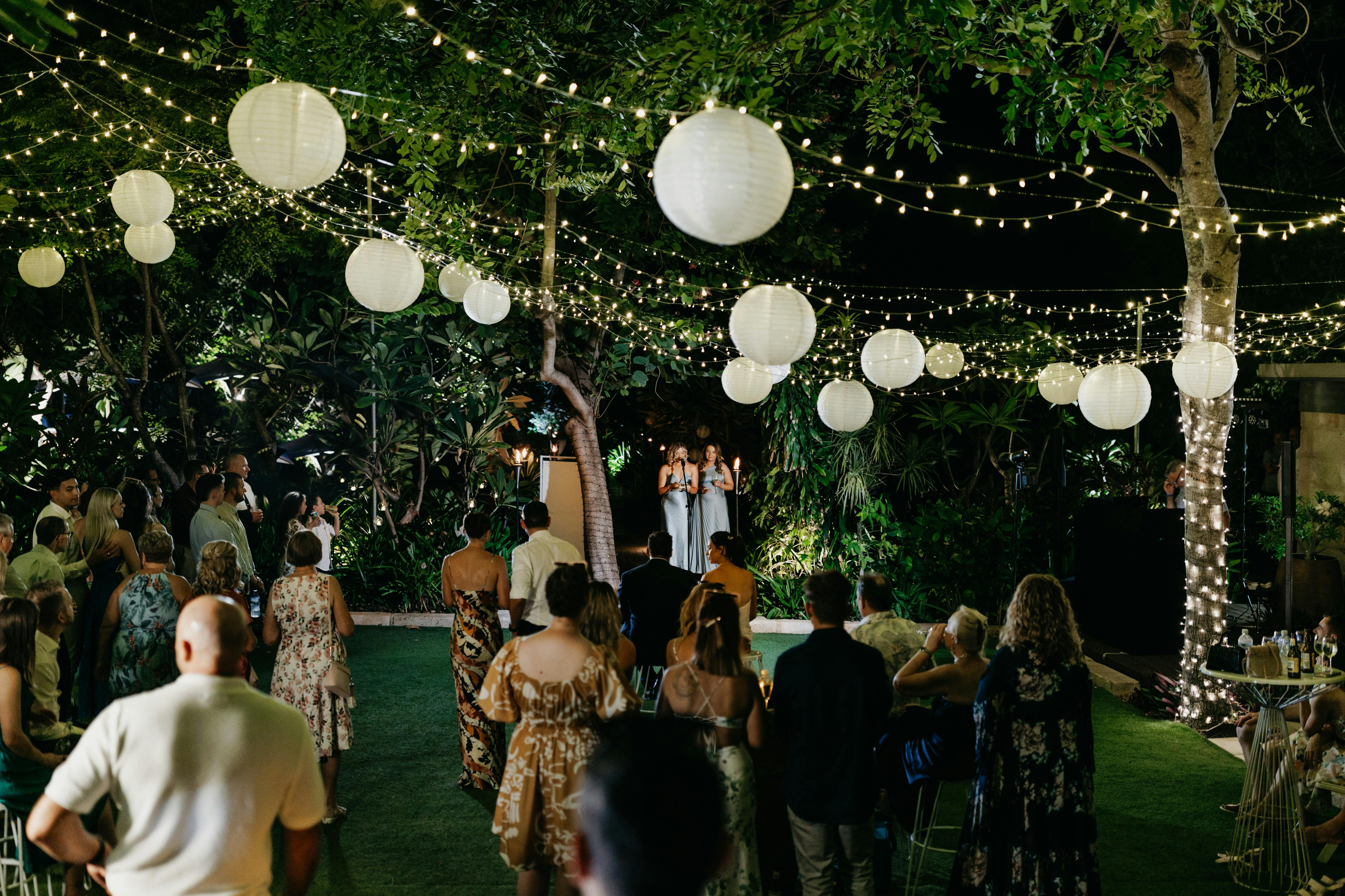 Guests enjoy speeches under fairy lights and lanterns at a magical outdoor wedding reception.