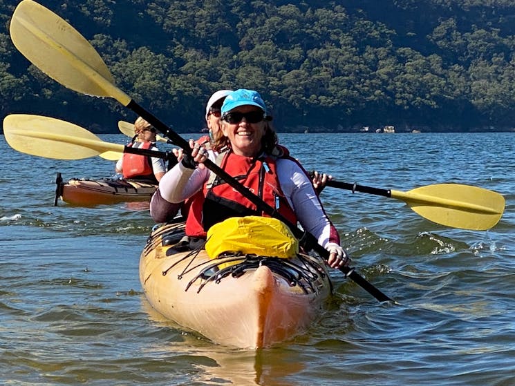 Two women on a yellow kayak wearing red life jackets, teal hat, yellow paddles in the air