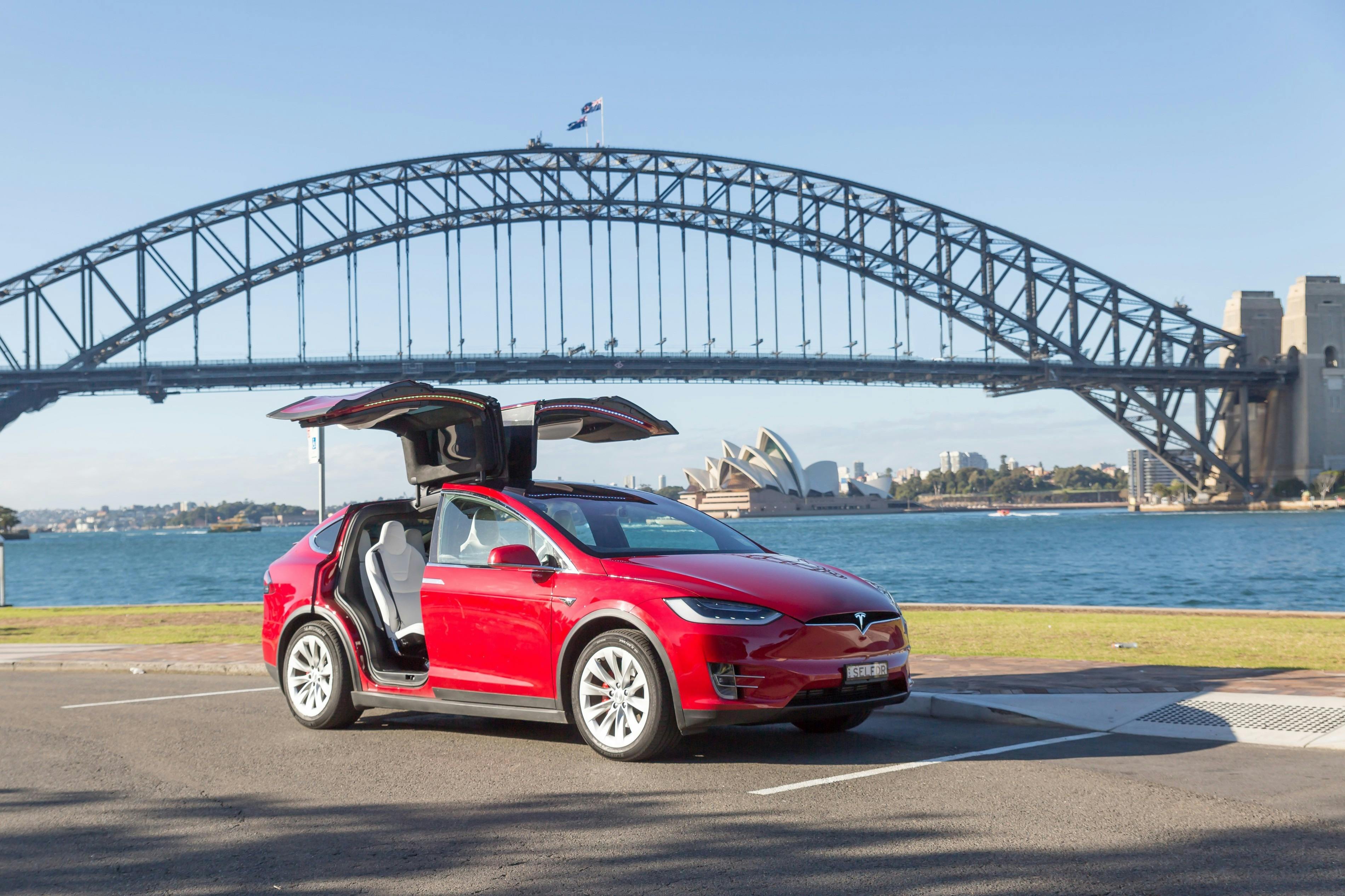 Red tesla parked below harbour bridge with wing doors open