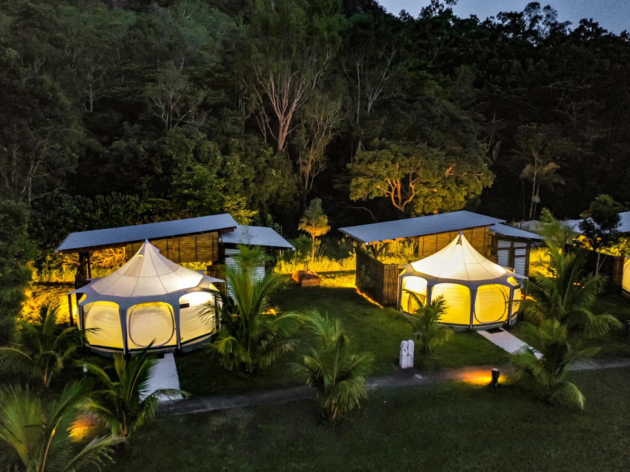 Warmly lit glamping tents surrounded by trees at night at Mt Jukes Eco Retreat. Warmly lit glamping tents surrounded by trees at night at Mt Jukes Eco Retreat.