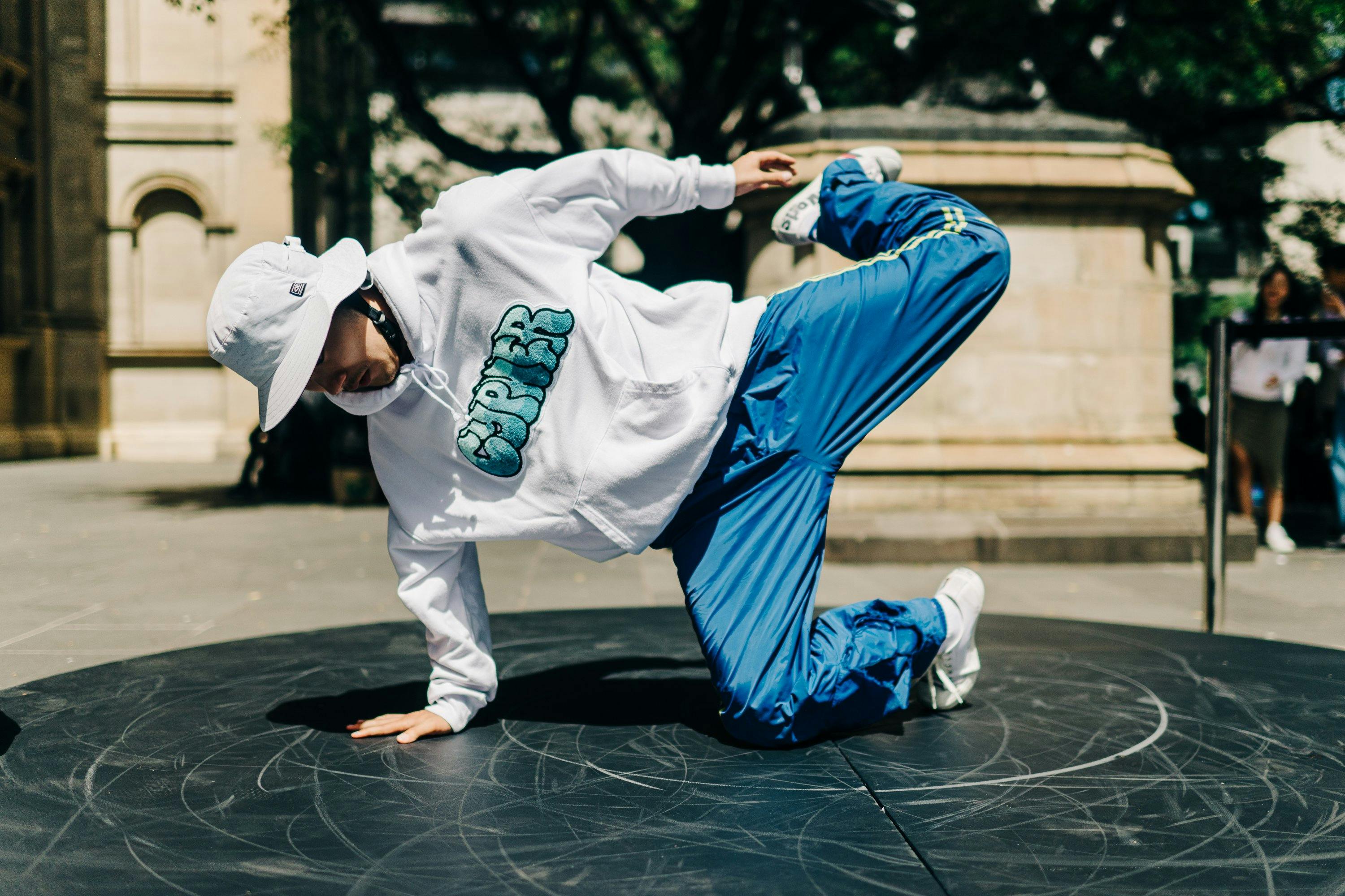 Dancer in blue pants, white jumper and white hat. They are mid movement, on a small circle stage.