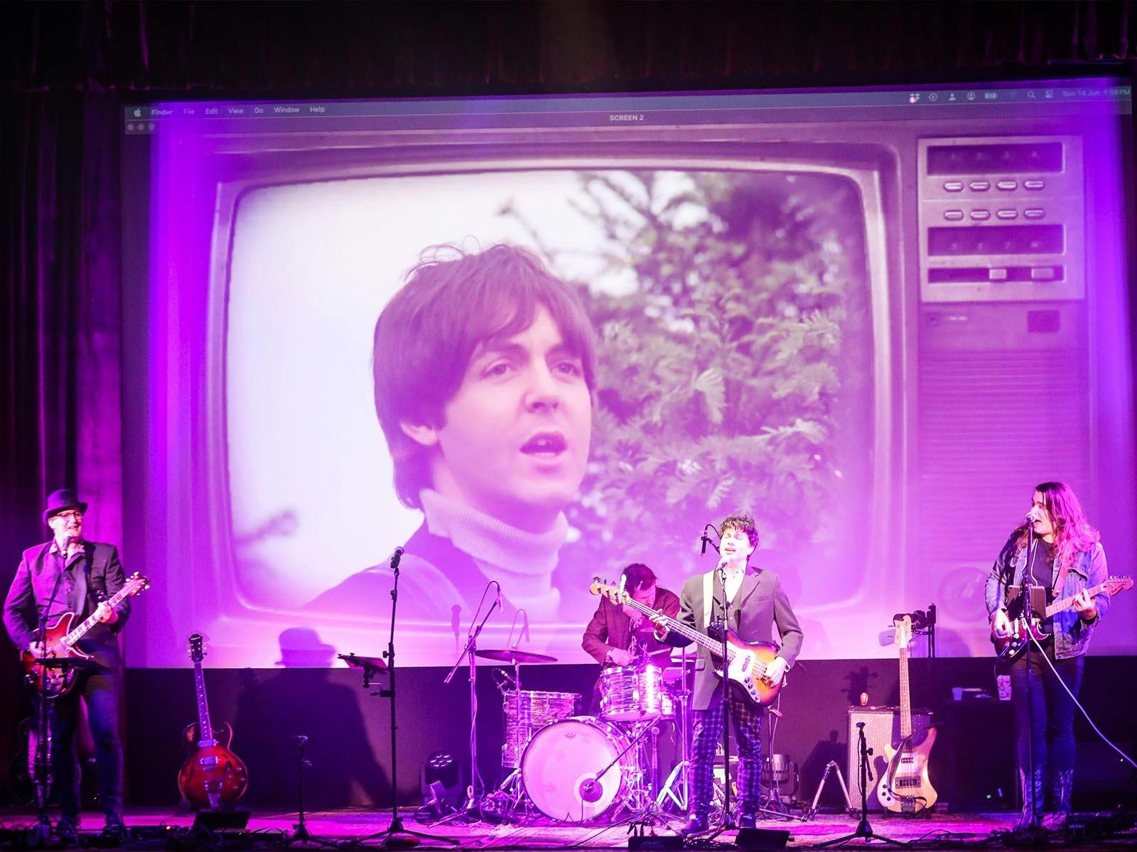 An acoustic band playing in front of a screen ft. a photo of Paul McCartney