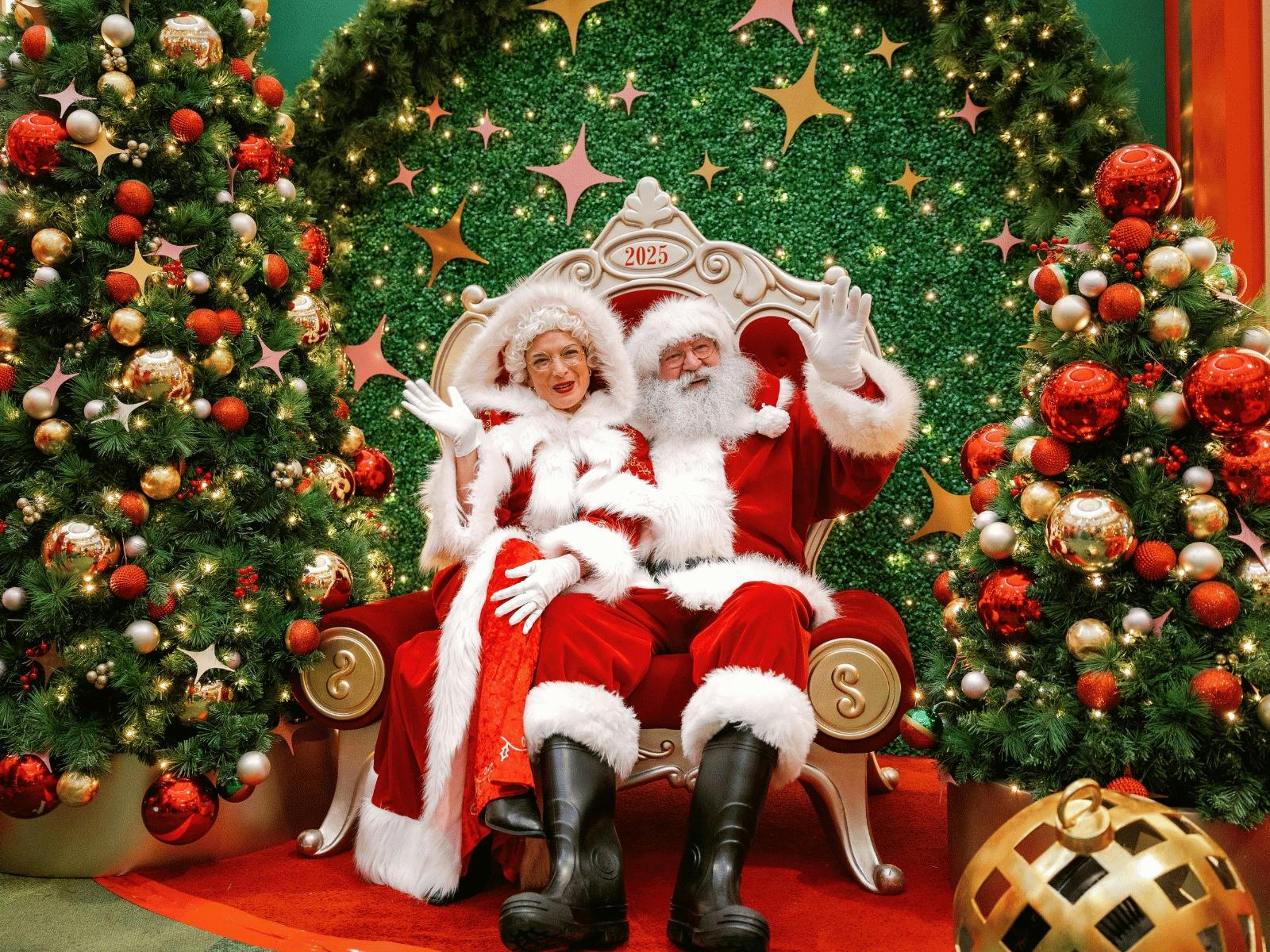 Santa Claus and Mrs Claus sitting in a read chair infront of a Christmas set waving.