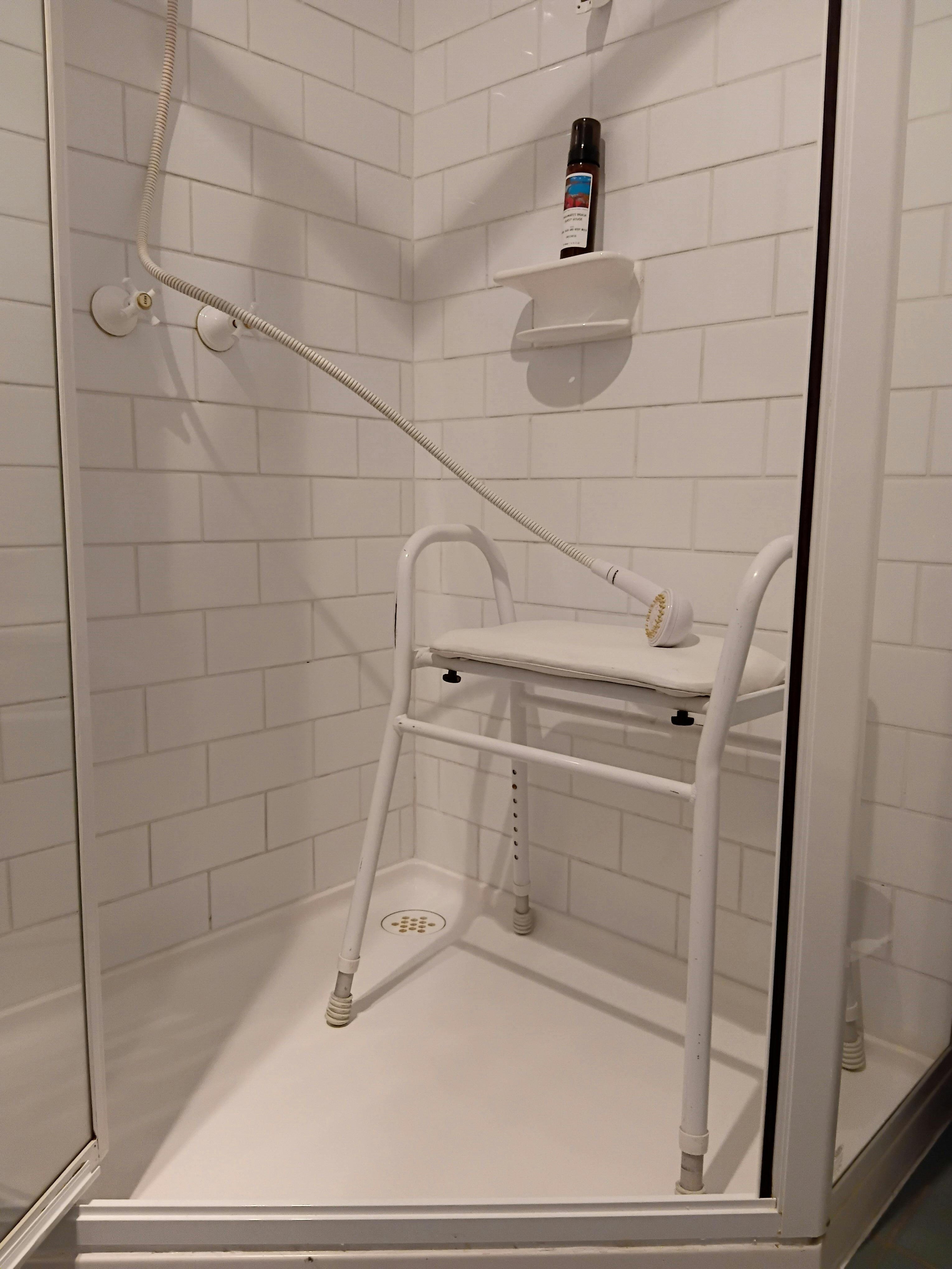 Padded shower seat standing on adjustable legs within a white tiled shower space.
