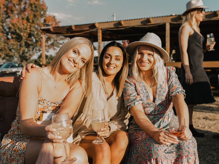 3 females drinking wine in the Autumn sun wearing dresses and hats