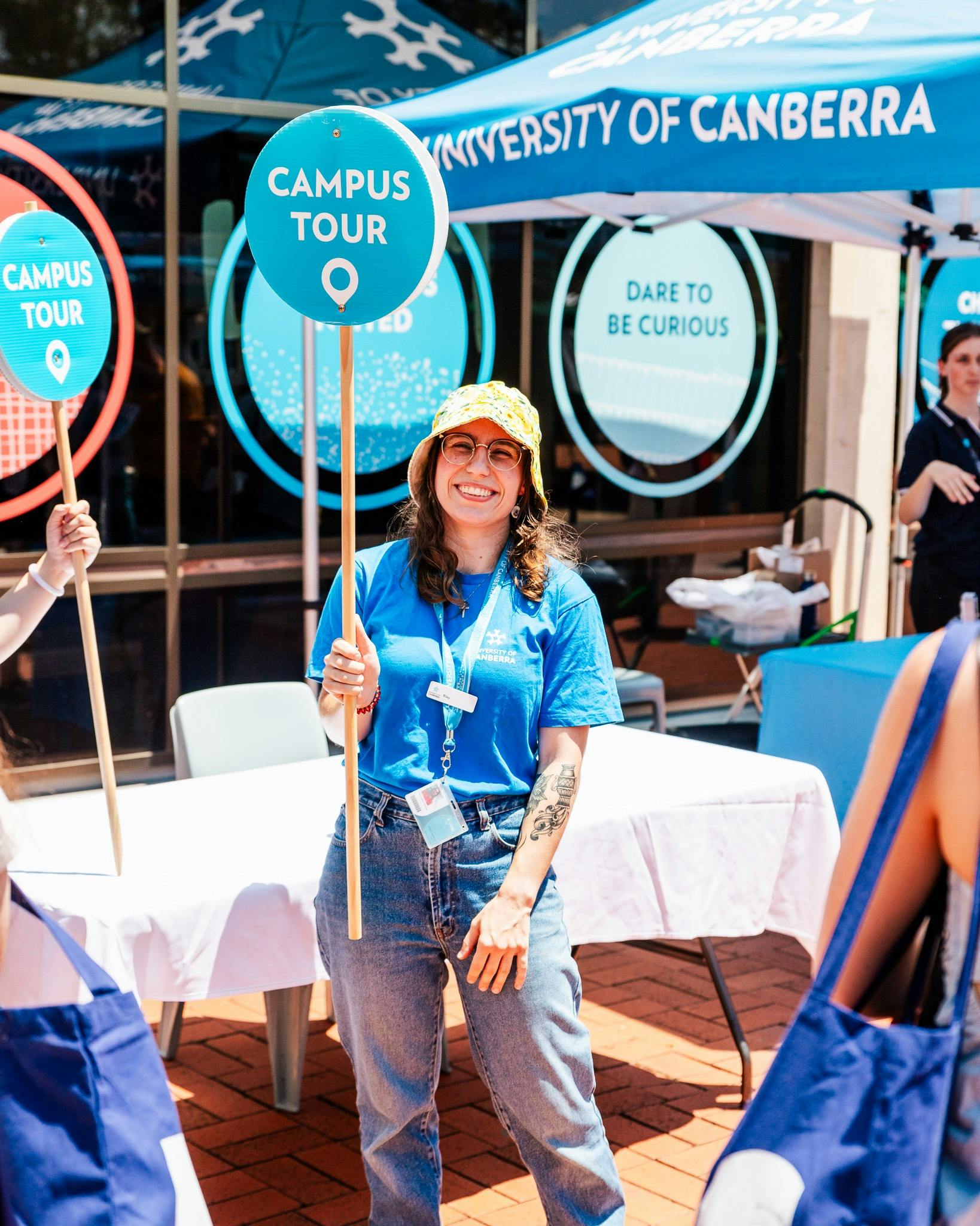 Happy UC staff member in blue shirt, holding a blue campus tours sign