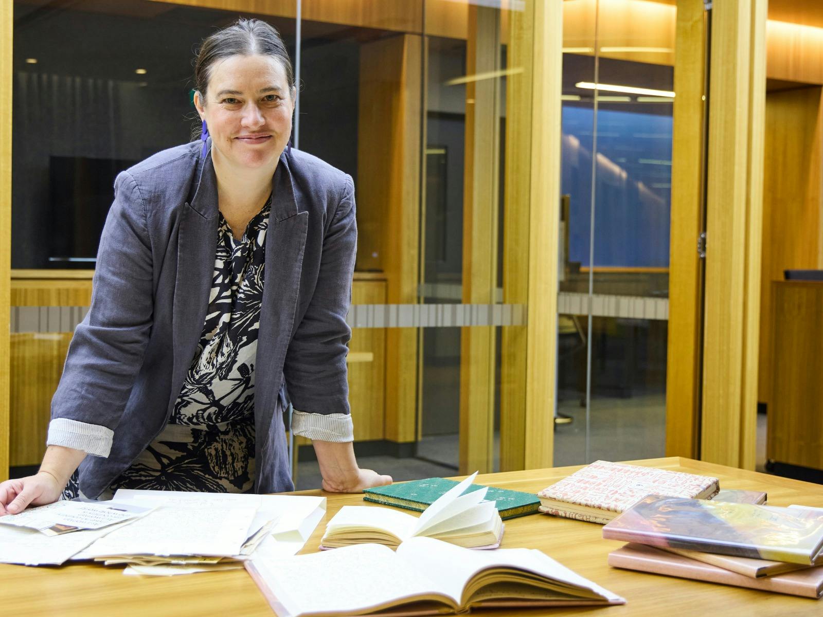 A woman in a grey blazer smiles at the camera while leaning on a wooden table covered with books.