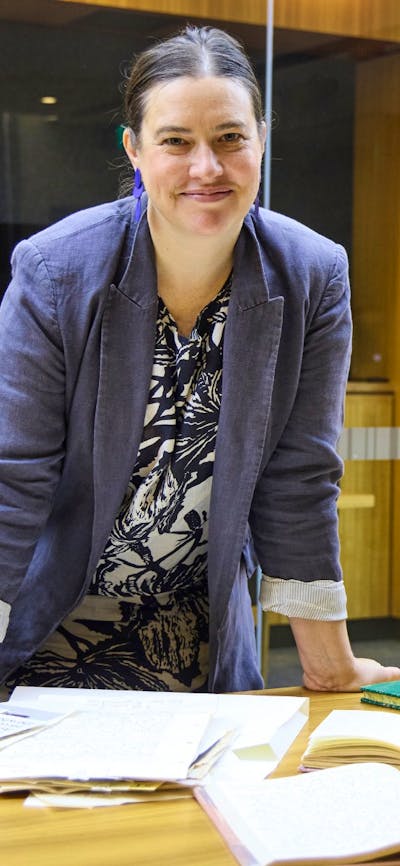 A woman in a grey blazer smiles at the camera while leaning on a wooden table covered with books.