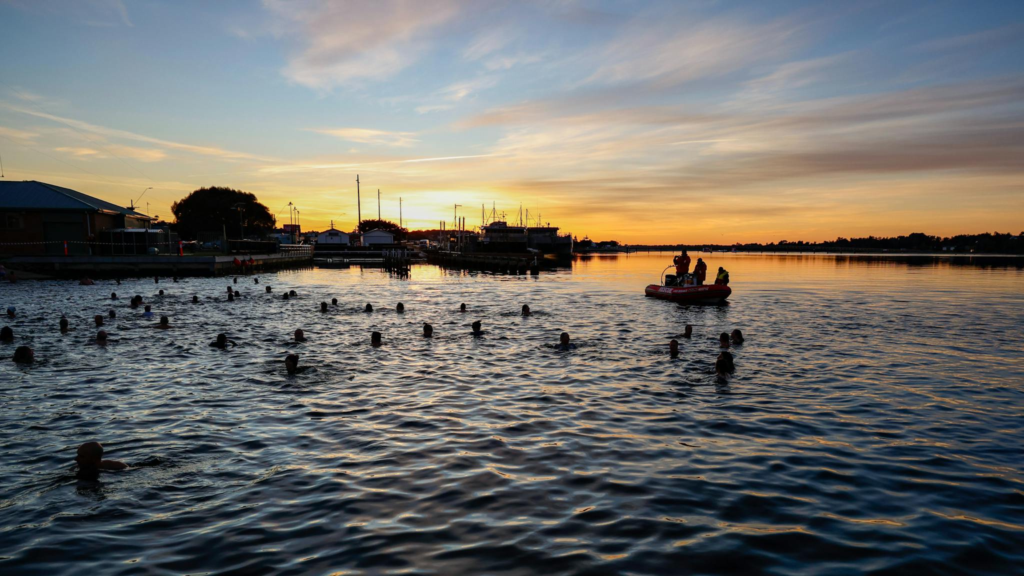 A large group of people swimming in a lake at sunrise with a coast guard boat in the background