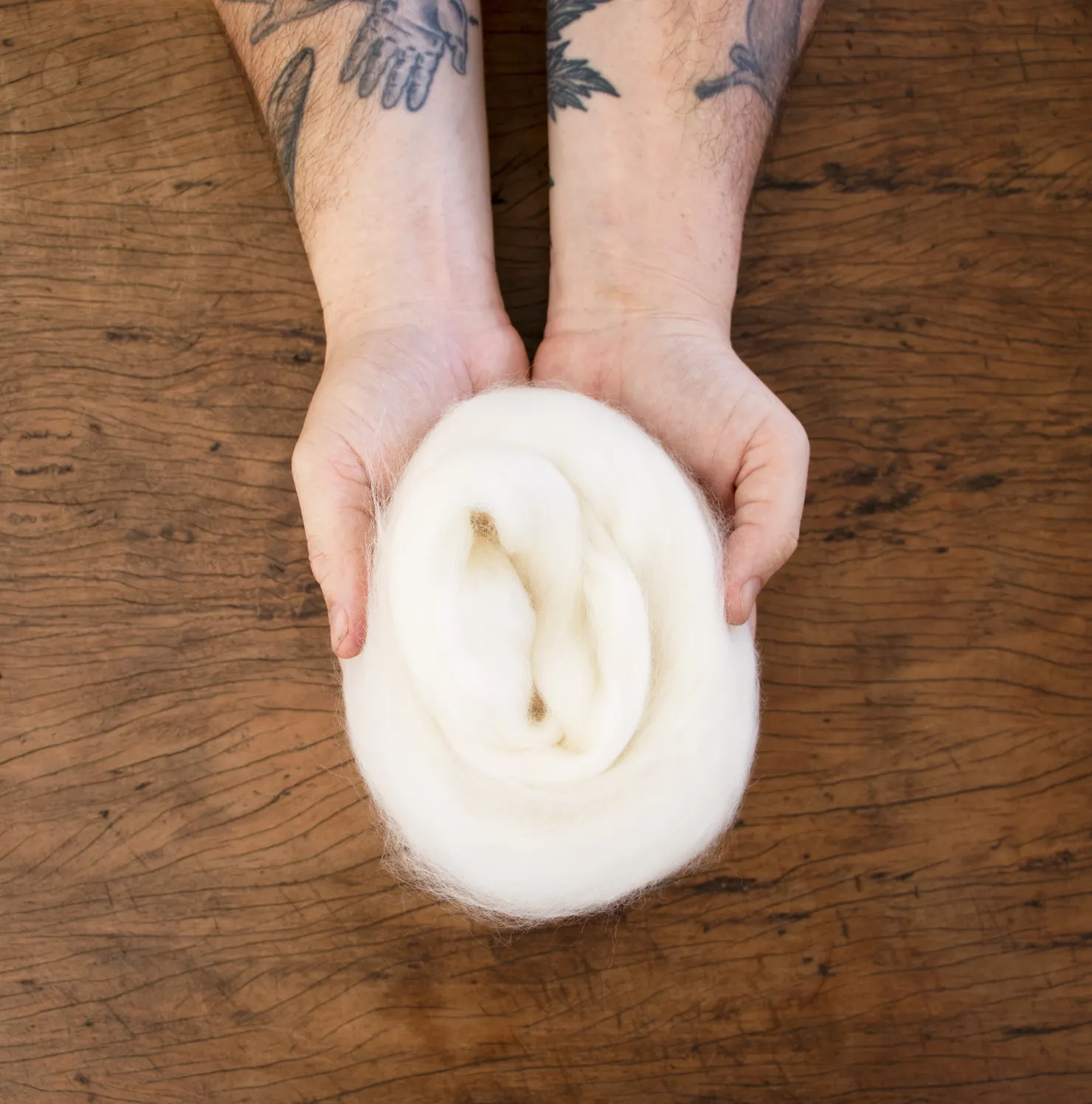 Two hands hold pale natural wool over a timber table top
