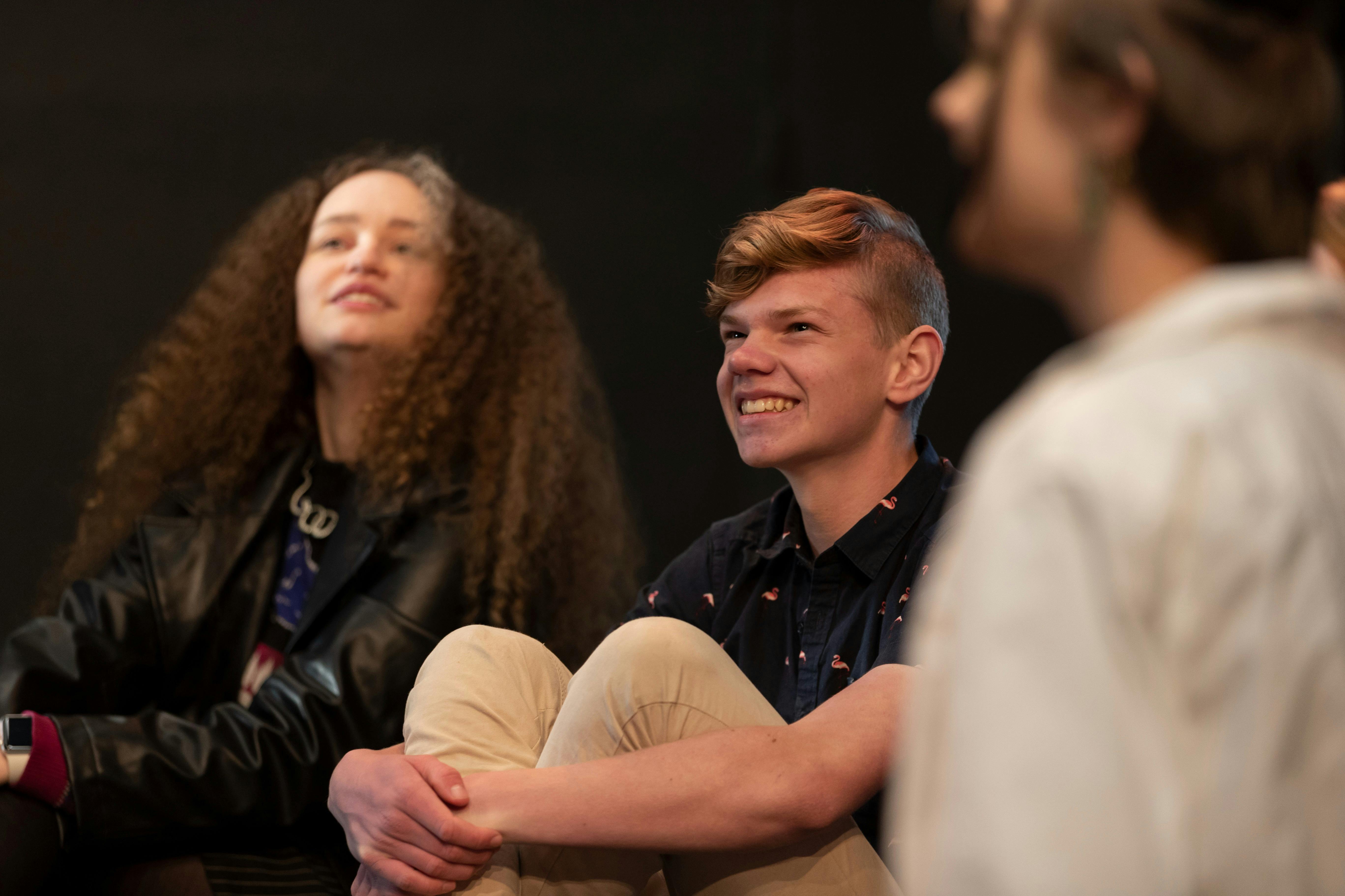 Three teenagers sit together in a theatre workshop, smiling as they enjoy watching a performance.