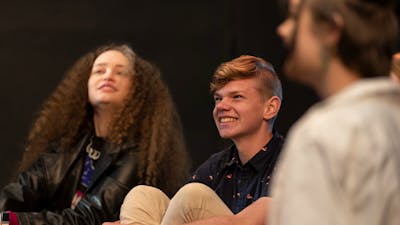 Three teenagers sit together in a theatre workshop, smiling as they enjoy watching a performance.