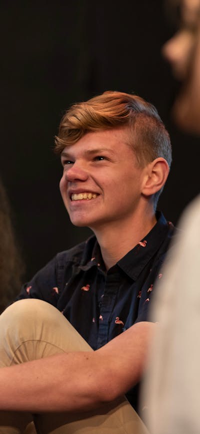 Three teenagers sit together in a theatre workshop, smiling as they enjoy watching a performance.
