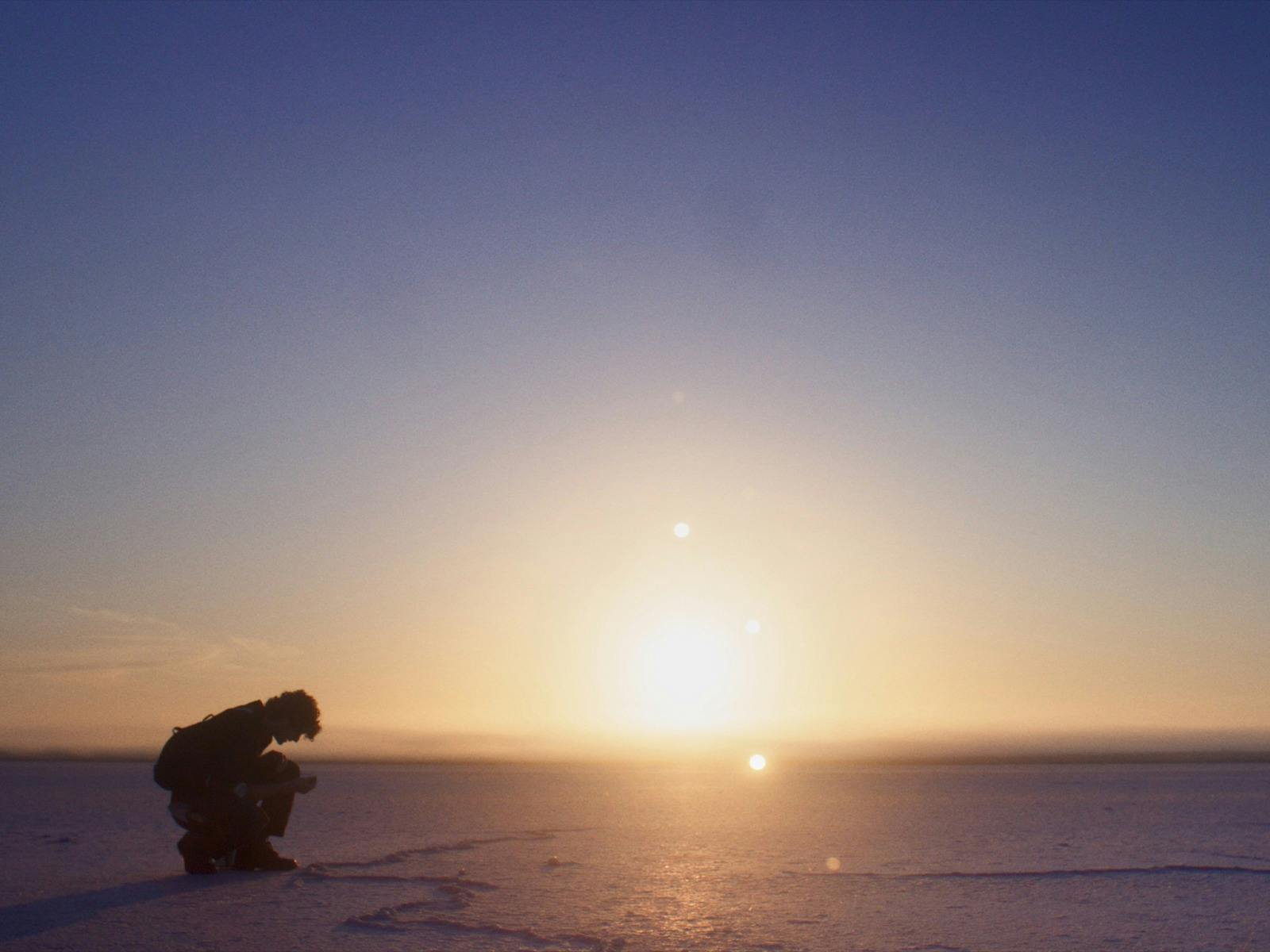 Silhouetted person (Josef) crouching on a snowy landscape at sunset.